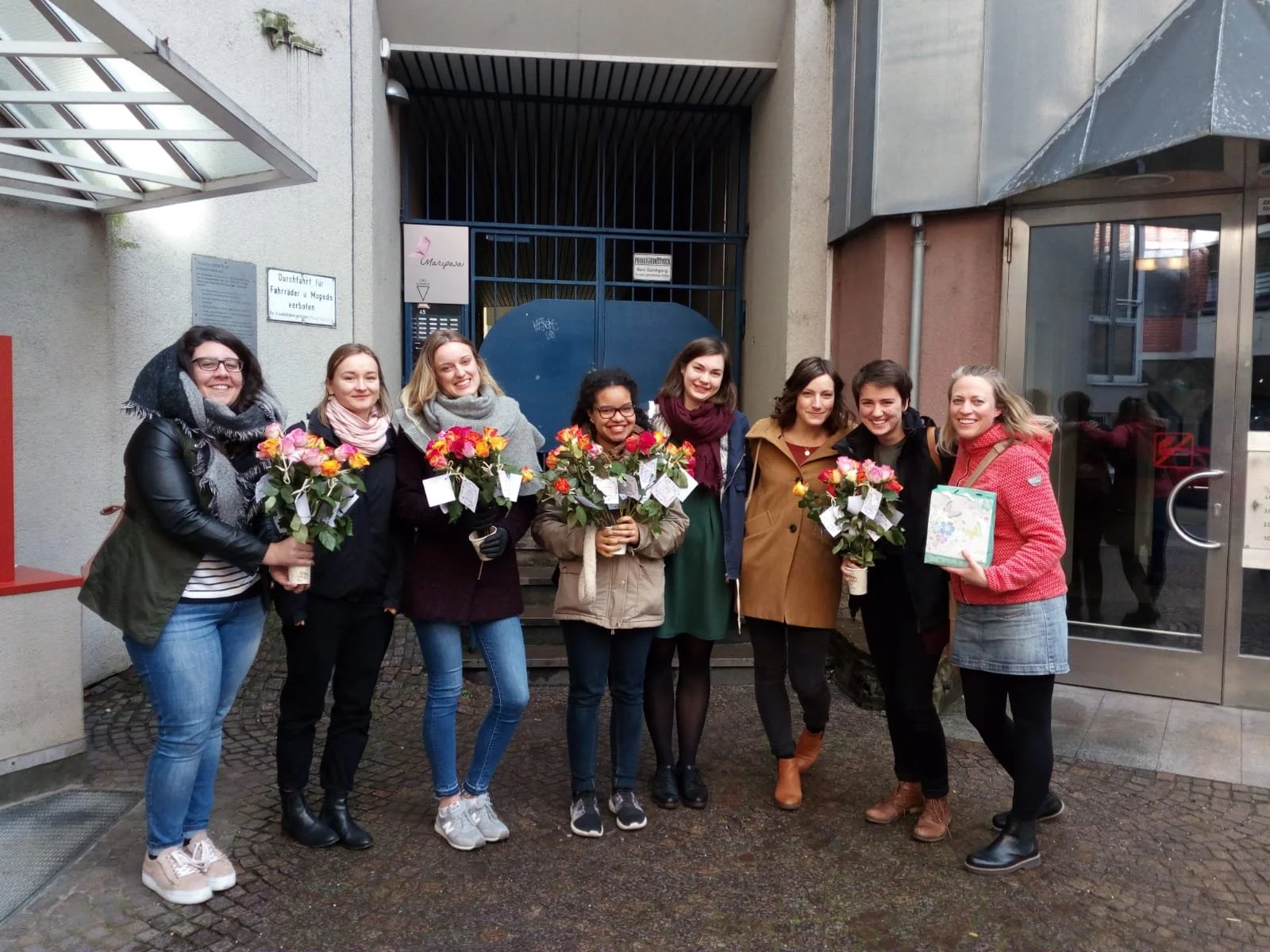 Eine Gruppe von acht Frauen steht draußen vor einem Gebäude, hält bunte Rosensträuße und eine Geschenkbox und lächelt in die Kamera.