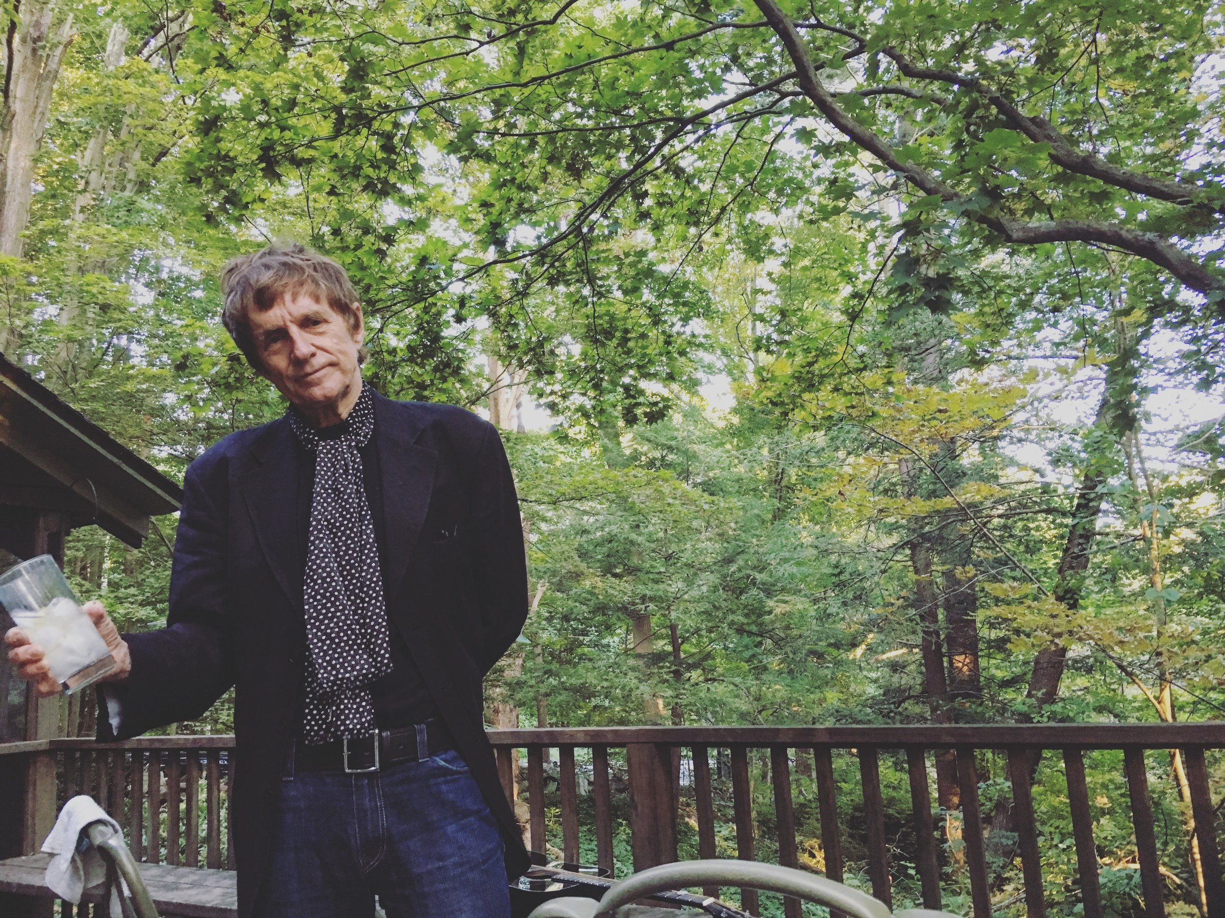 the artist stands among the trees holding a cocktail and seeming to say goodbye