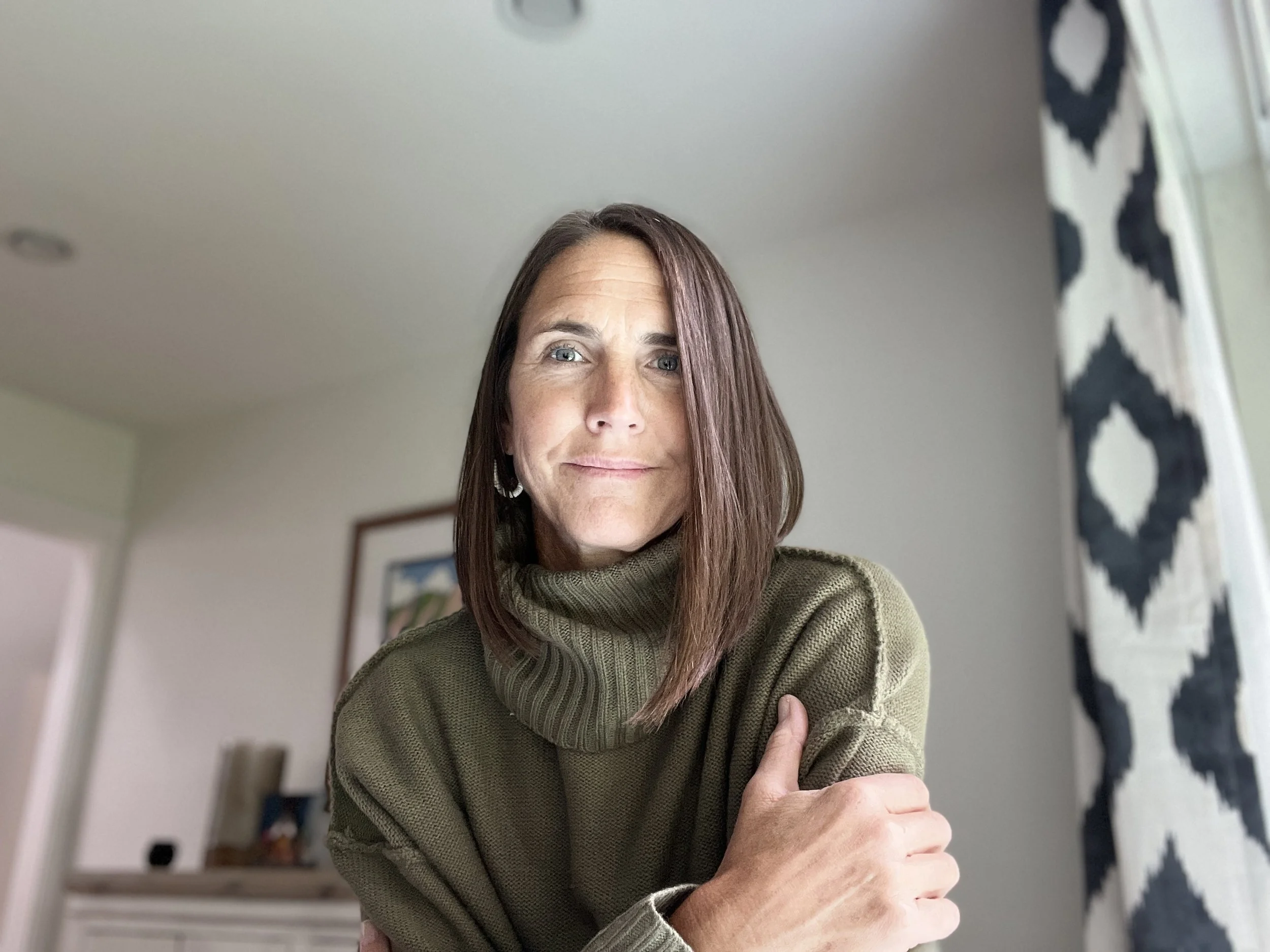 A woman with shoulder-length brown hair wearing an olive green turtleneck sweater poses indoors.