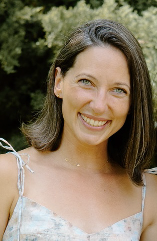 A woman with shoulder-length dark hair smiling outdoors, with trees in the background.