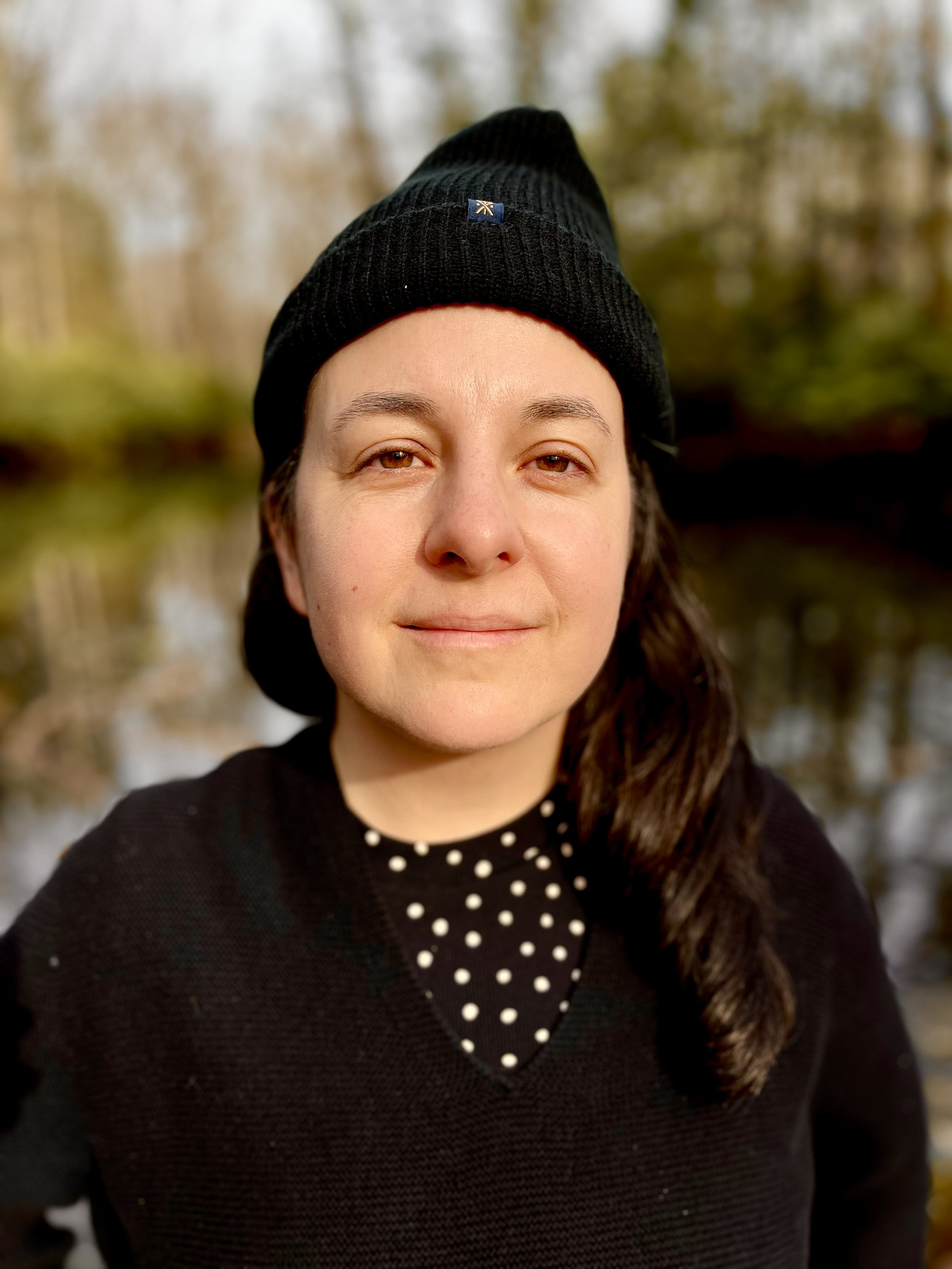 A woman wearing a black beanie and black sweater with white polka dots, standing outdoors near a body of water with trees in the background.