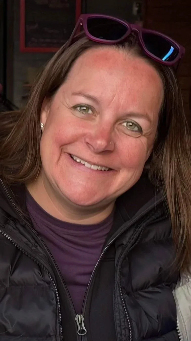 Close-up of a smiling woman with brown hair, wearing purple sunglasses on her head, earrings, a nose piercing, a purple shirt, and a black puffer jacket.