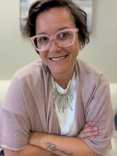 A woman with short brown hair, wearing pink glasses, smiling, sitting indoors with arms crossed, in front of a neutral background.