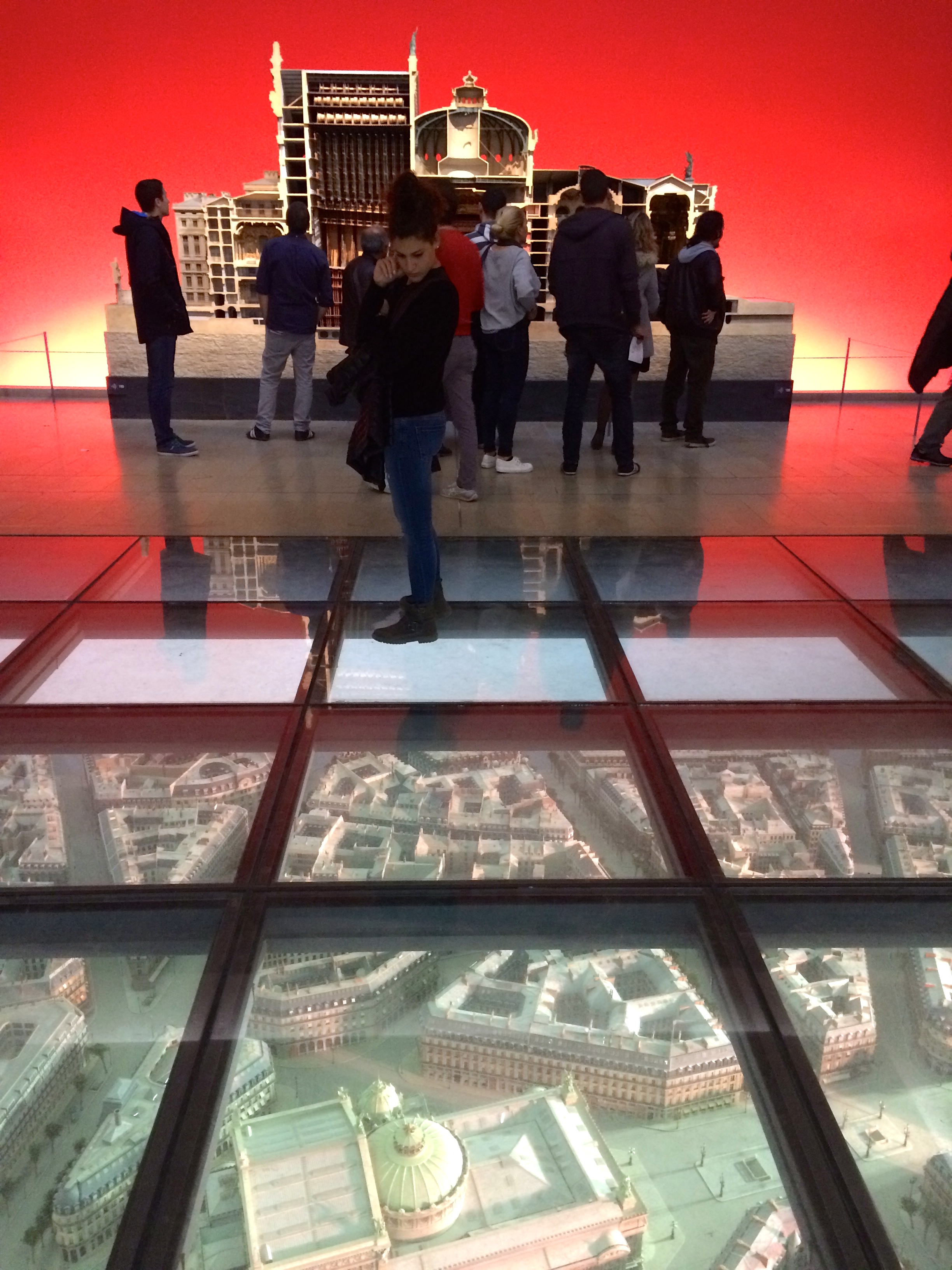 Visitors peer downward at a large model of the Opera neighbourhood beneath a glass floor for context.