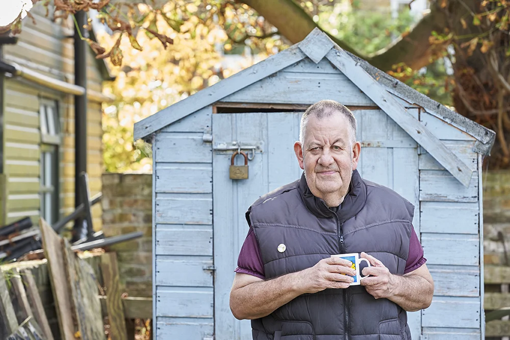 Photograph of an older man with a cup of tea stood in front of his shed