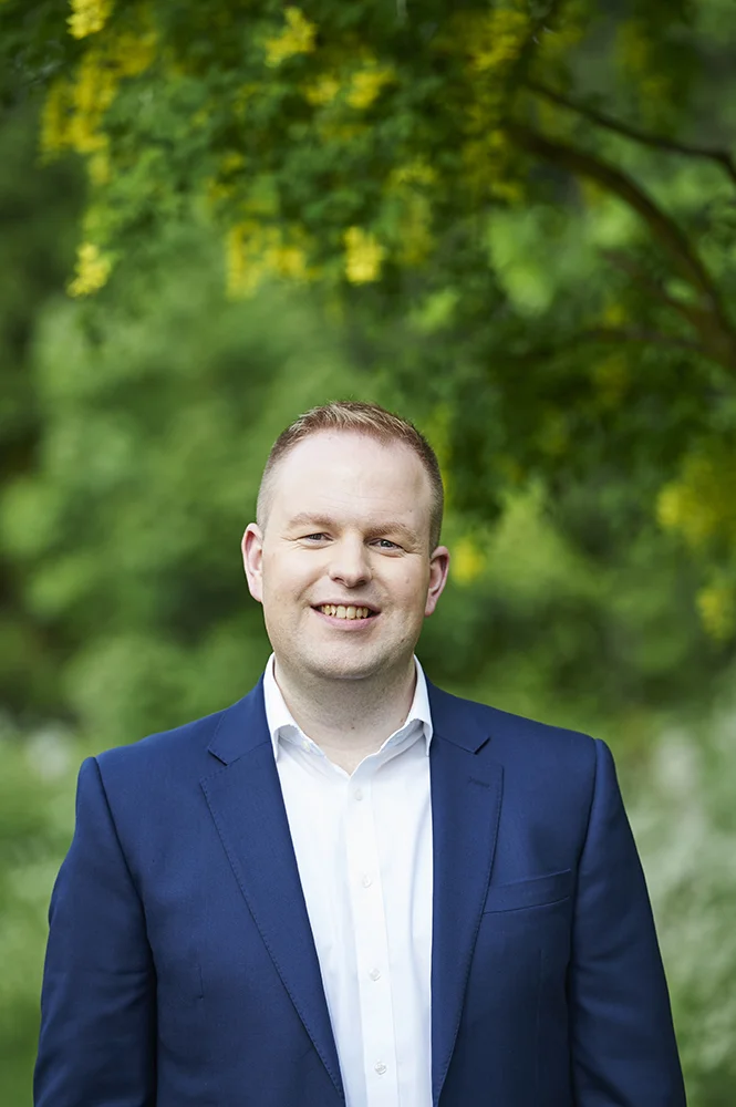 Smiling Headshot Photo of a man in a blue suit, stood under a tree