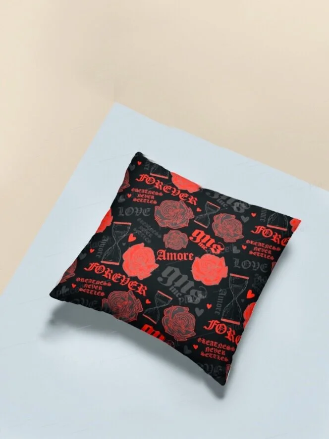 A square pillow with a black background decorated with red and pink roses, hearts, and French words like 'Amore' and 'Forte,' placed on a white surface against a light-colored wall.