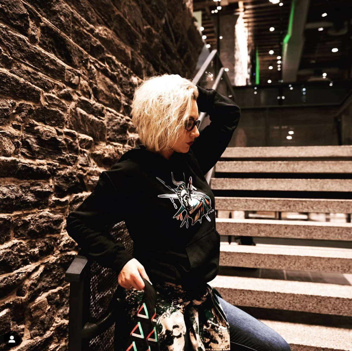Woman with curly blonde hair and glasses, sitting on a black chair against a stone wall and stairs, wearing a black hoodie with a graphic of a unicorn and colorful patterned pants, in an indoor setting with warm lighting.