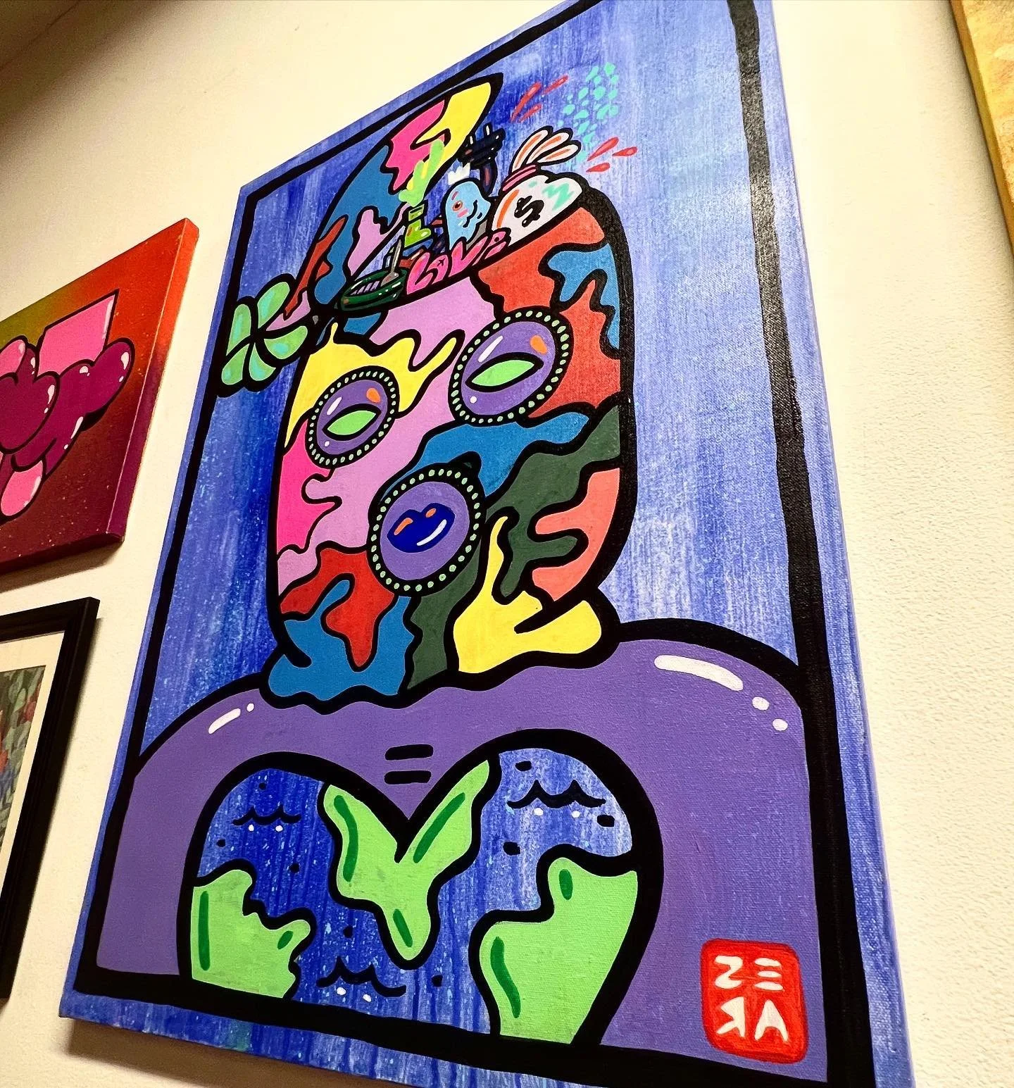 Colorful abstract art hanging on a wall, featuring a stylized face with large, dotted eyes and a melting appearance, above a depiction of Earth with landmasses and water, and a red artist's signature in the bottom right corner.