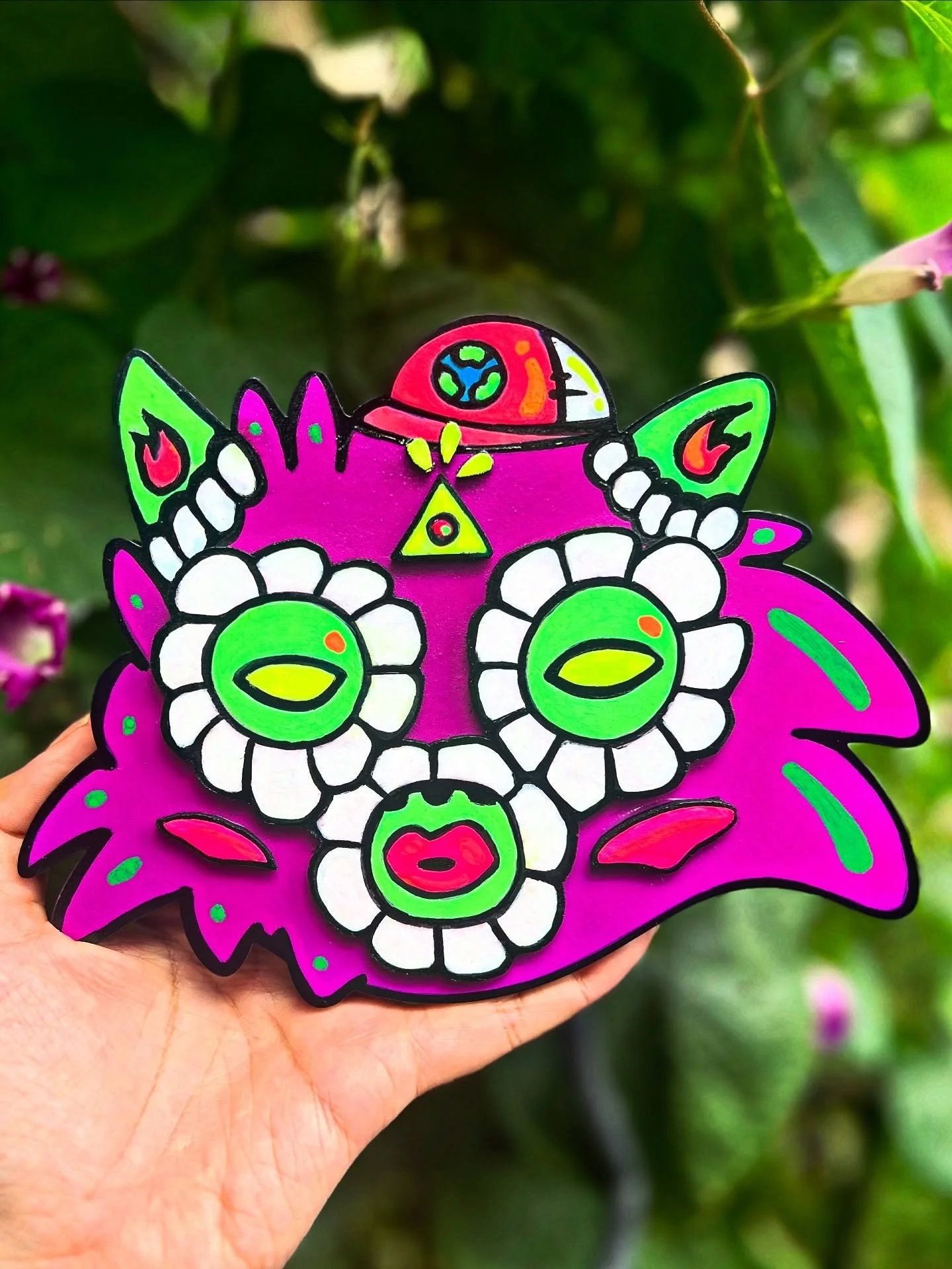 Colorful decorative mask with vibrant pink, green, white, and red colors, featuring floral and abstract designs, held in hand against a leafy background.