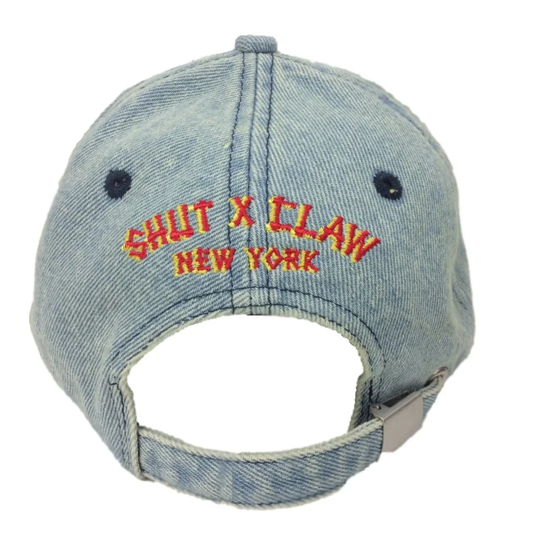Back of a light blue denim baseball cap with embroidered red and yellow text reading 'SHUT X CLAN NEW YORK' and small black eyelets.