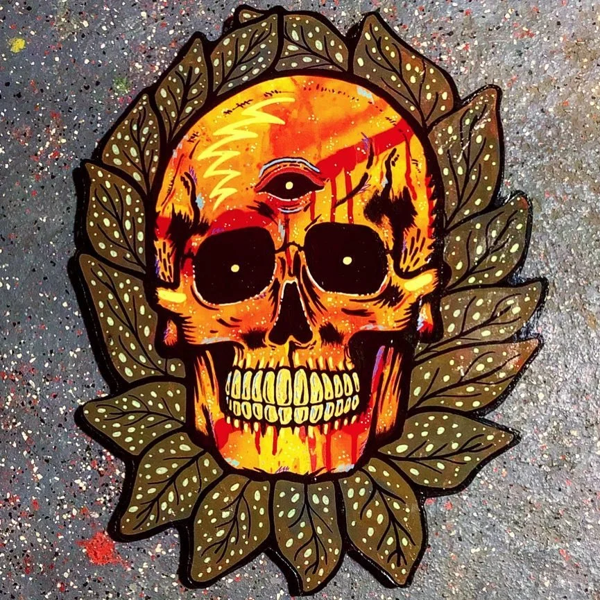 A colorful street art mural of a skull with an eye on its forehead, surrounded by green leaves with white spots, on a speckled concrete surface.