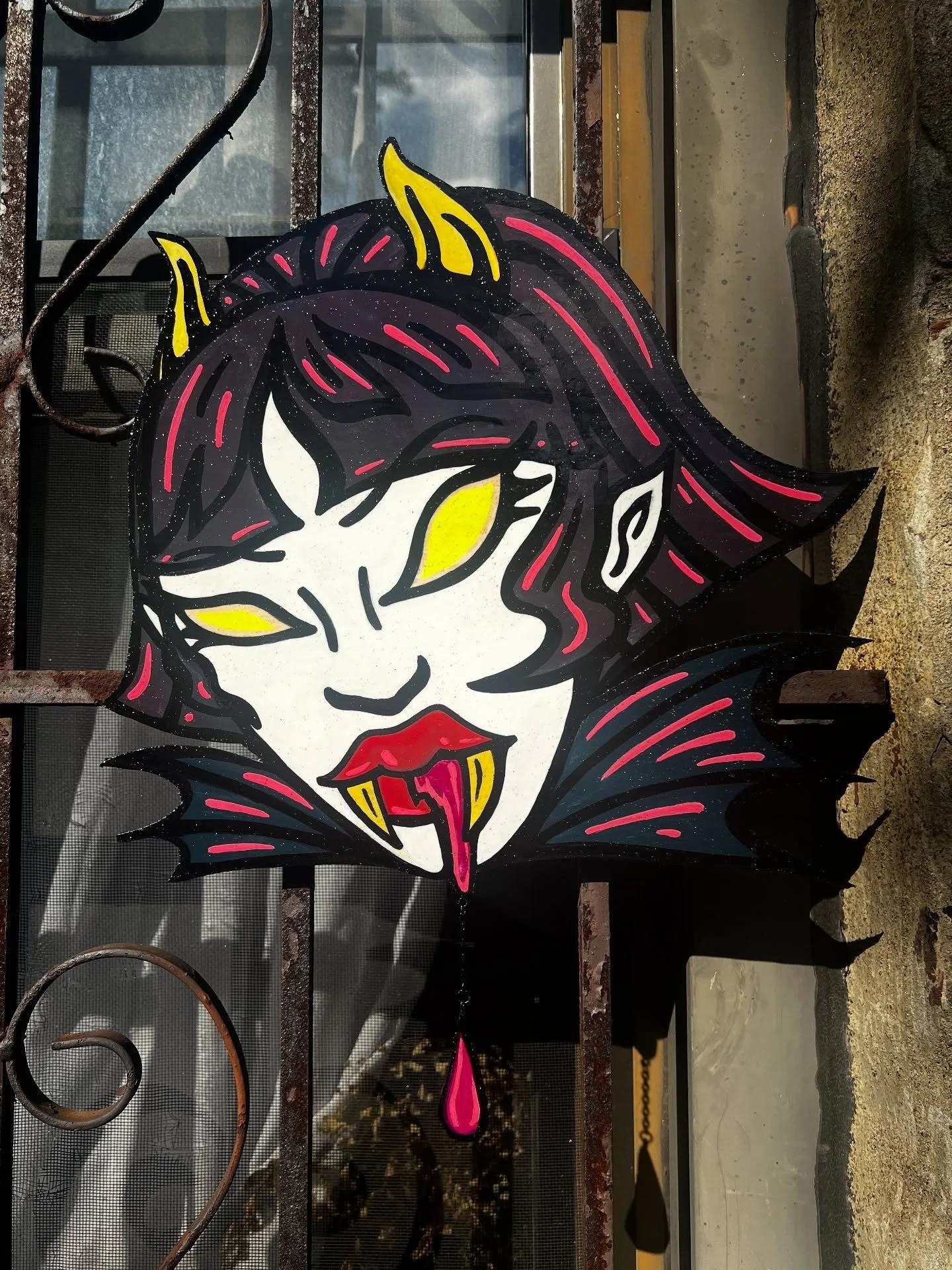 A decorative Halloween-themed sign of a vampire girl with yellow eyes, black and red hair, fangs, and dripping blood, mounted on a metal fence.