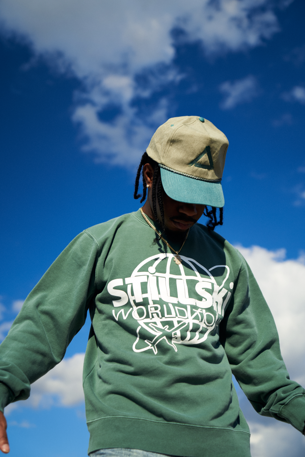 A person wearing a green sweatshirt with white graphics and text, a beige and teal cap, and a chain necklace, standing outdoors against a partly cloudy blue sky.