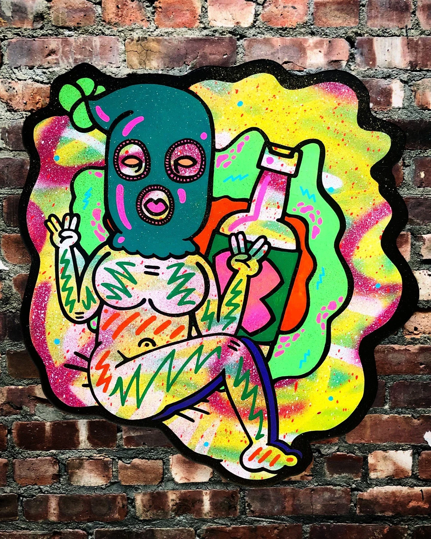 Colorful graffiti art of a person wearing a ski mask with a green bow, sitting relaxed with crossed legs, holding up peace signs with both hands, background features bright splashes of yellow, pink, green, and blue on a brick wall.