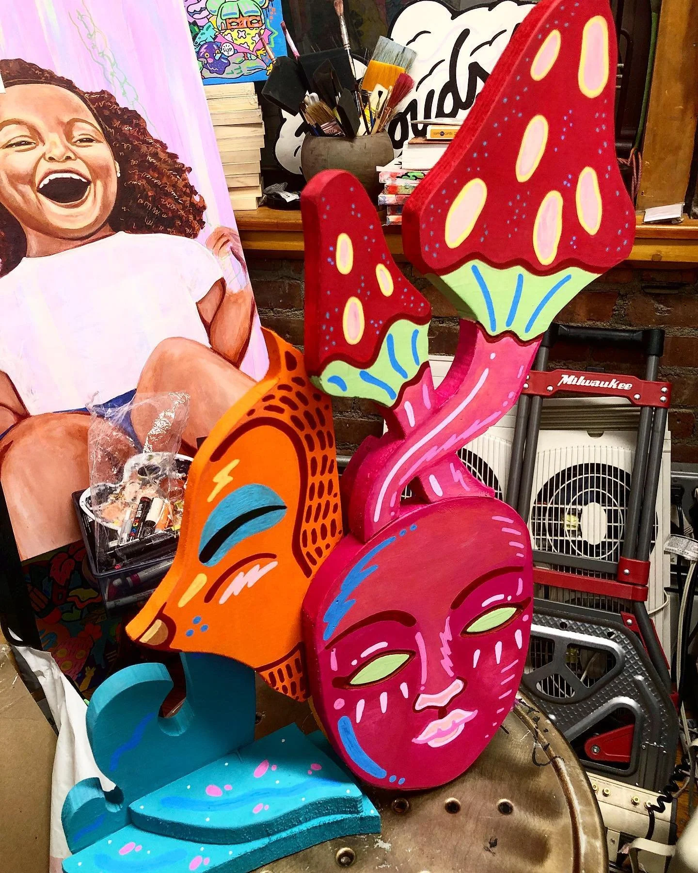 Colorful three-dimensional art sculptures of a pink face with closed eyes, blue eyebrows, and lips; an orange face with closed eyes and blue accent; and a red flower with yellow spots and a green base, all in a vibrant, playful style.