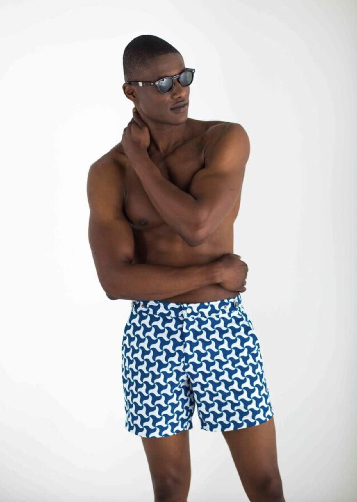 A young man with sunglasses, no shirt, and patterned swim shorts standing against a plain white background.