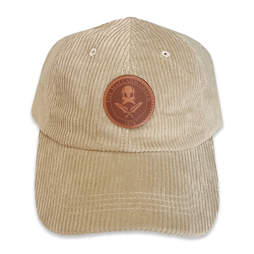 Beige corduroy baseball cap with a round brown leather patch featuring a skull and crossbones, surrounded by a circle with the text 'The Mallard Order 1576' and small eyelets on the crown.