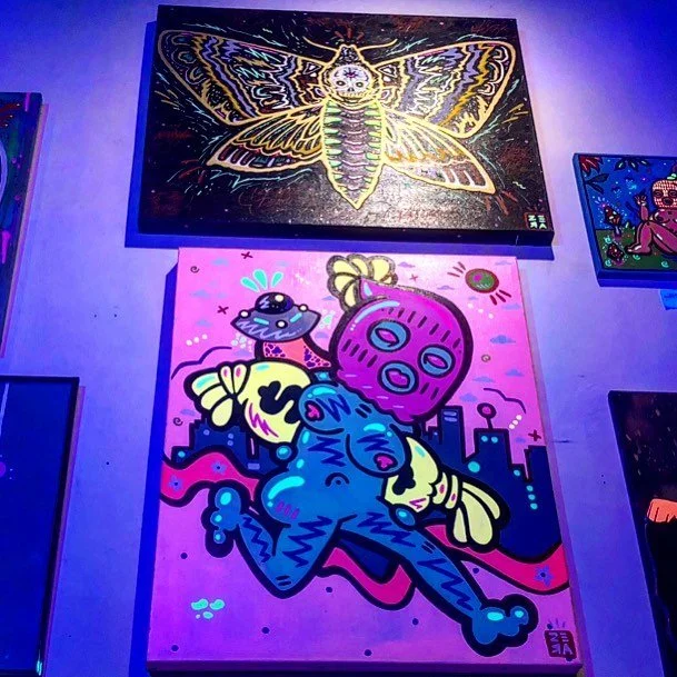 Colorful street art murals including a large butterfly with intricate yellow, purple, and black wings at the top, a pink face with multiple eyes and antennae floating above a blue creature with yellow wings, set against a cityscape background. Neon l