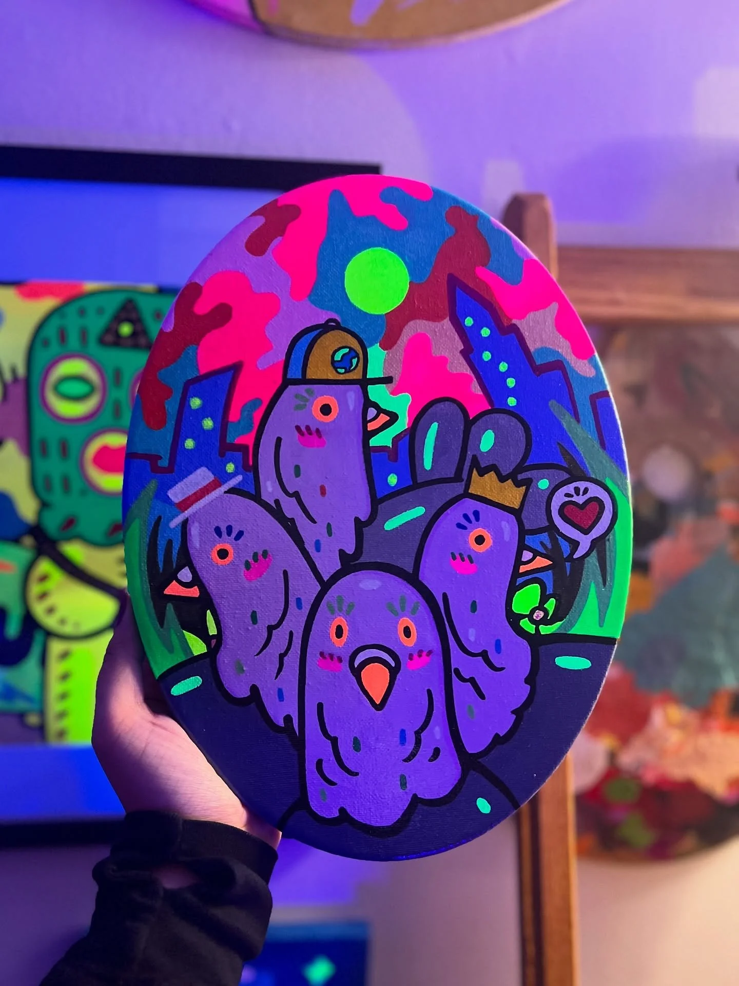 A hand holding a painted rock featuring neon-colored cartoon chickens, purple with orange beaks, set against a vibrant background with buildings, a green sun, and a small heart symbol.
