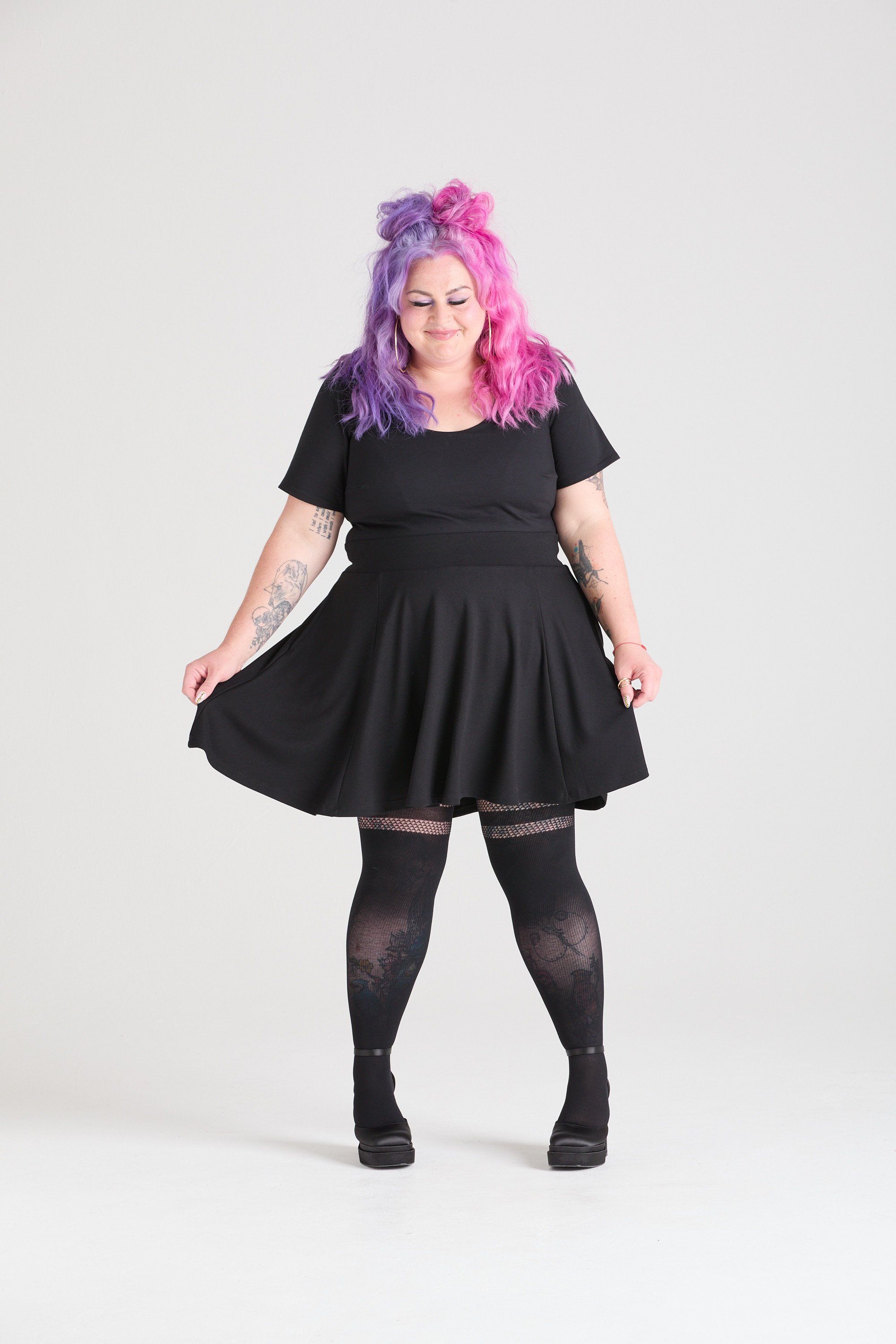 A woman with pink and purple curly hair, wearing a black dress, patterned tights, and high heels, standing against a plain light gray background.