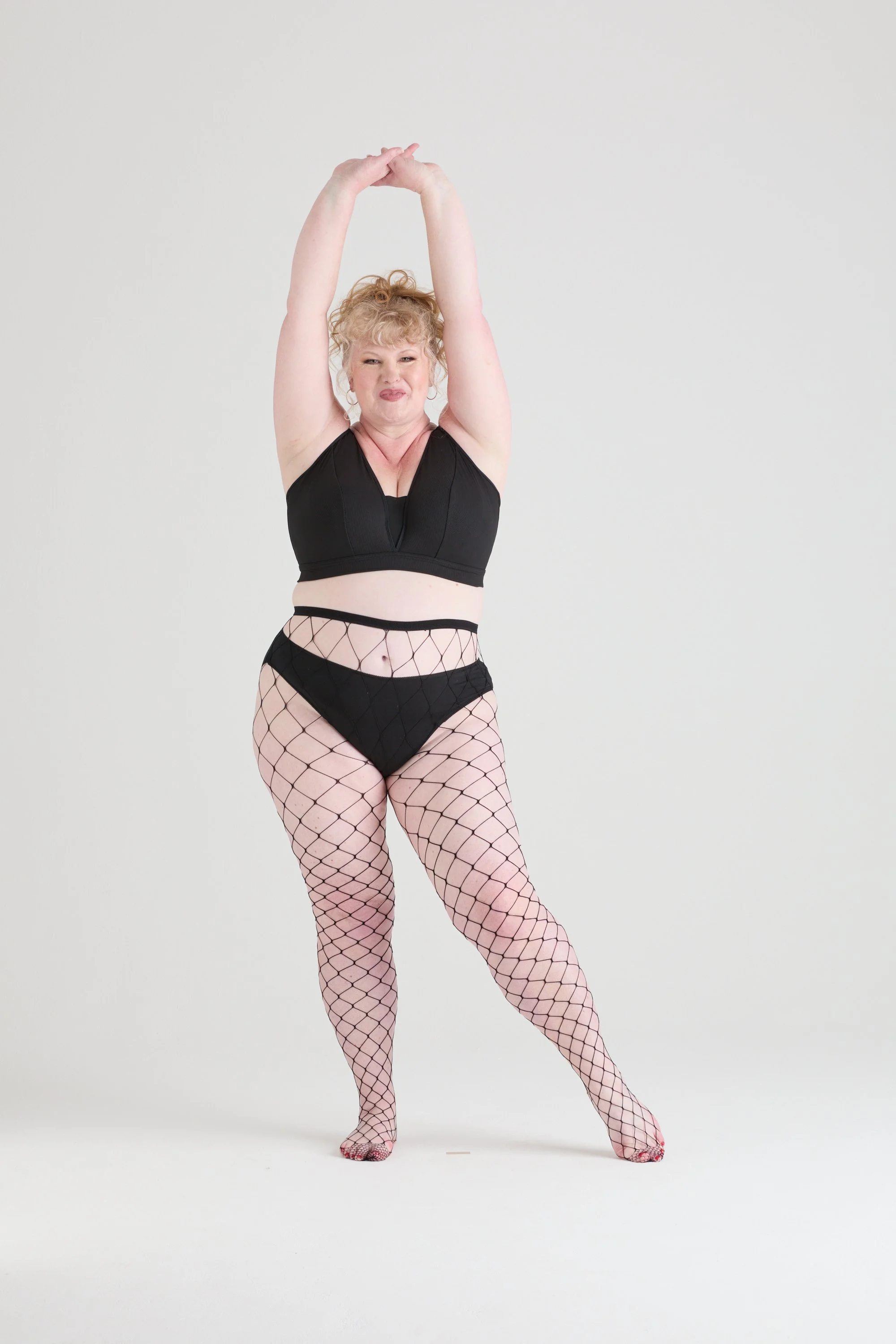 Woman with curly blonde hair in a black sports bra and fishnet tights striking a dance pose against a plain white background.