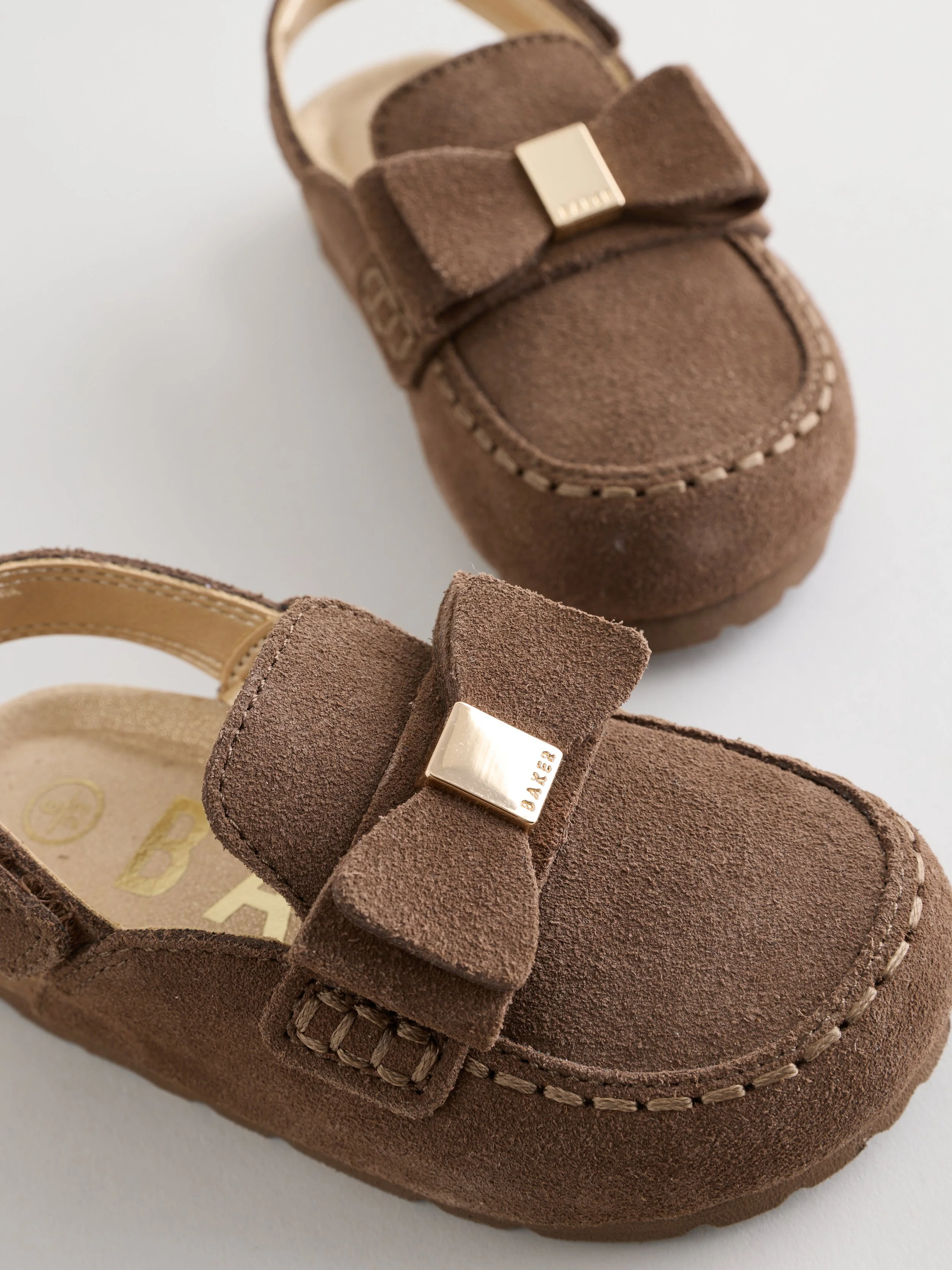 Close-up of a brown suede moccasin shoe with a decorative bow and metallic hardware on the instep, against a white background.