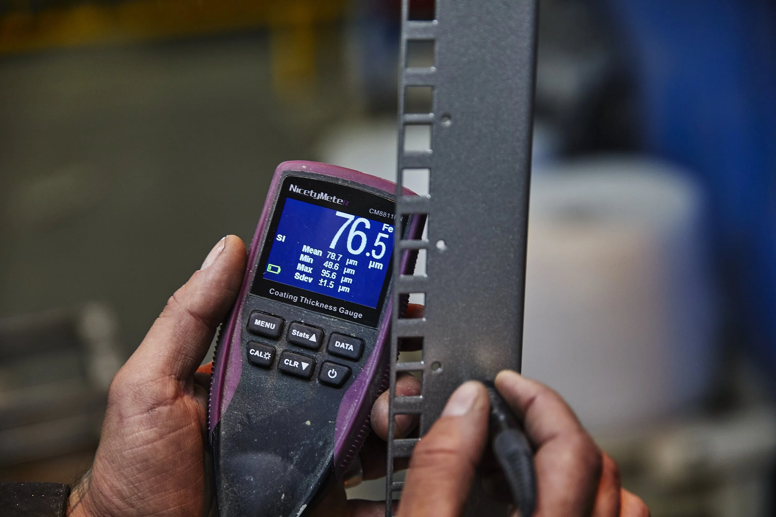 A person holding a pink NicetyMeter R coating thickness gauge measuring the thickness of a metal shelf or fixture.