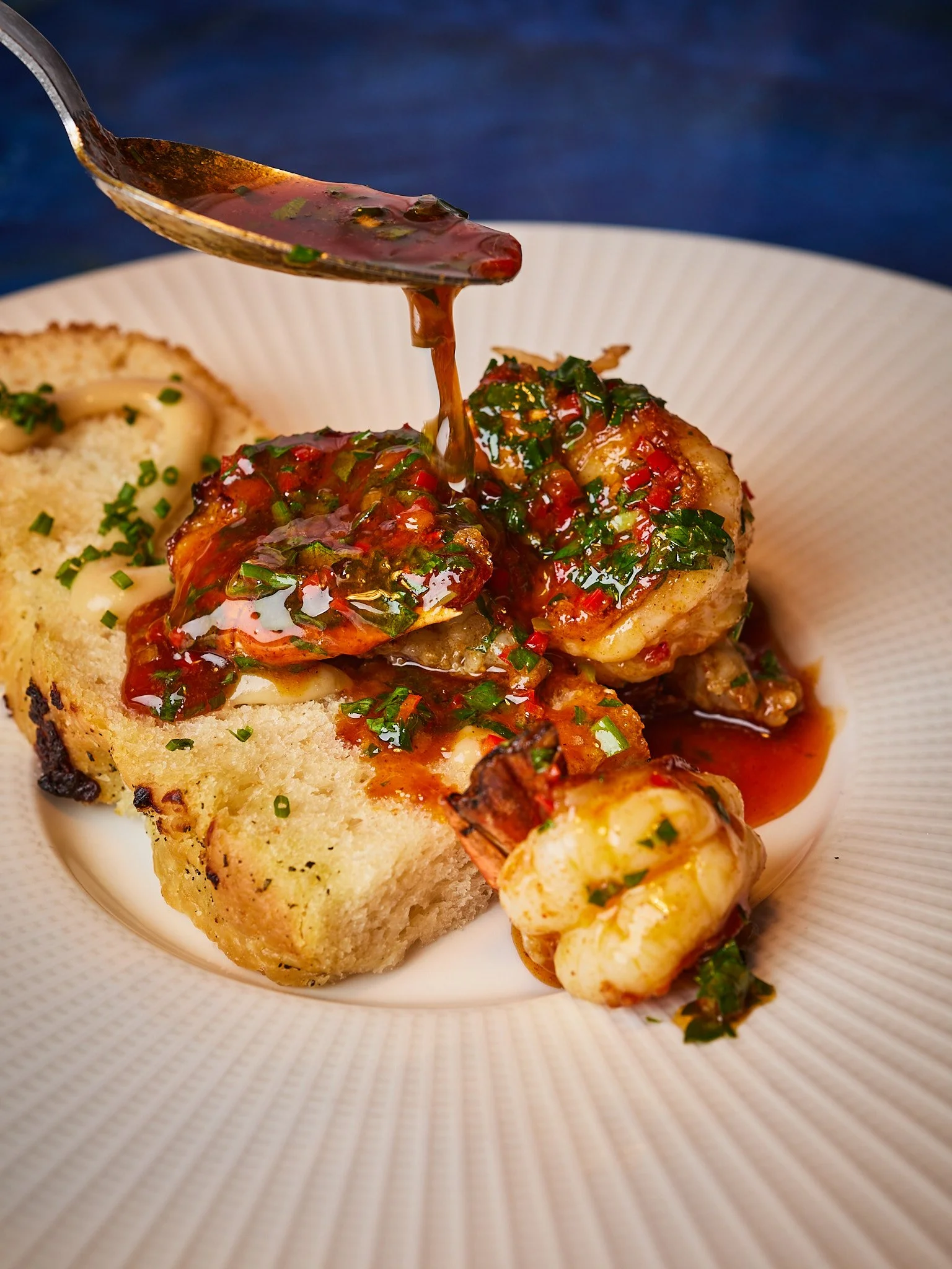 Close-up of a savory dish with cooked shrimp topped with a spicy tomato and herb sauce, served with a slice of bread on a white plate.