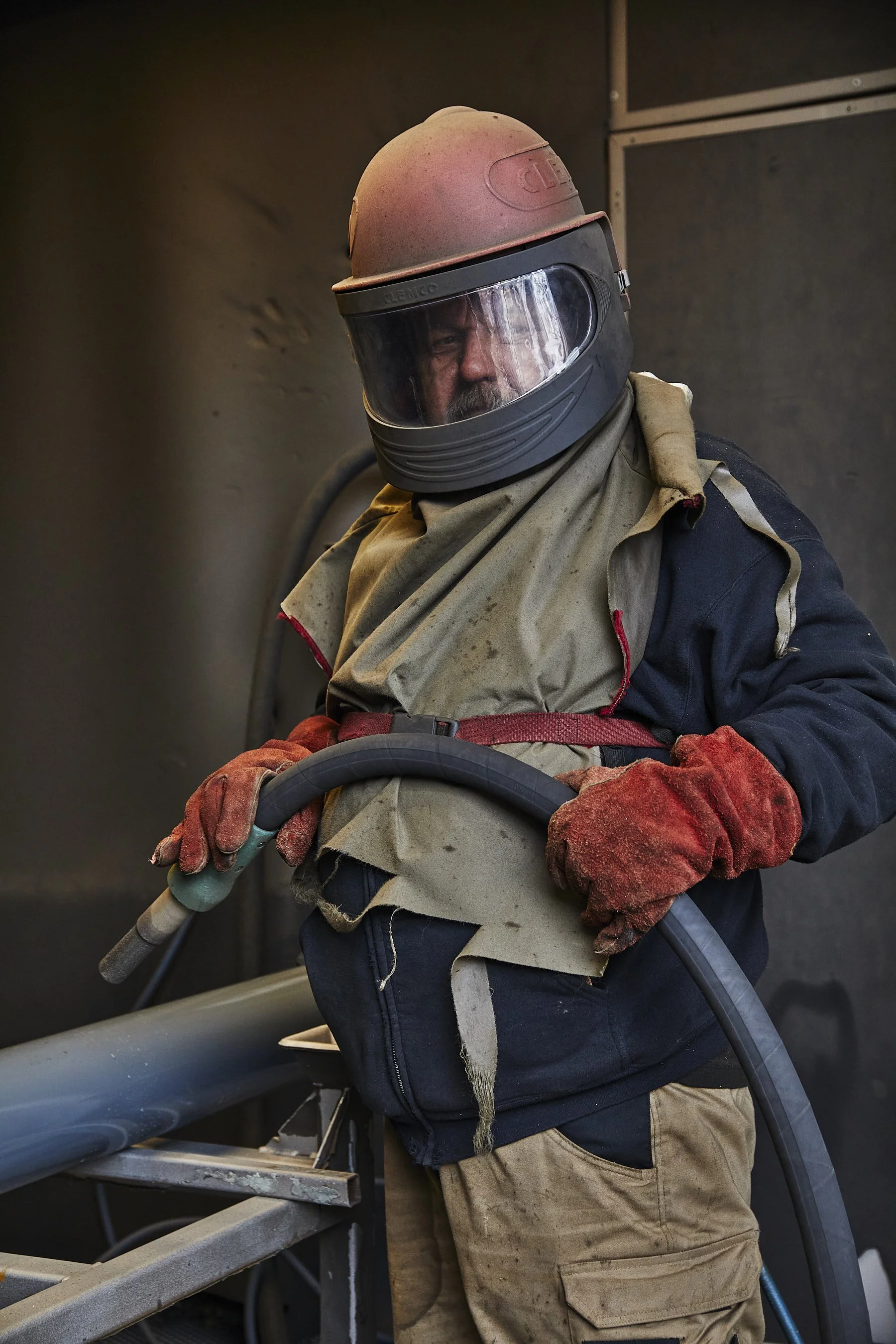 A construction worker wearing a beige hard hat, safety goggles, red gloves, and a beige apron is working with a large hose on a construction site.