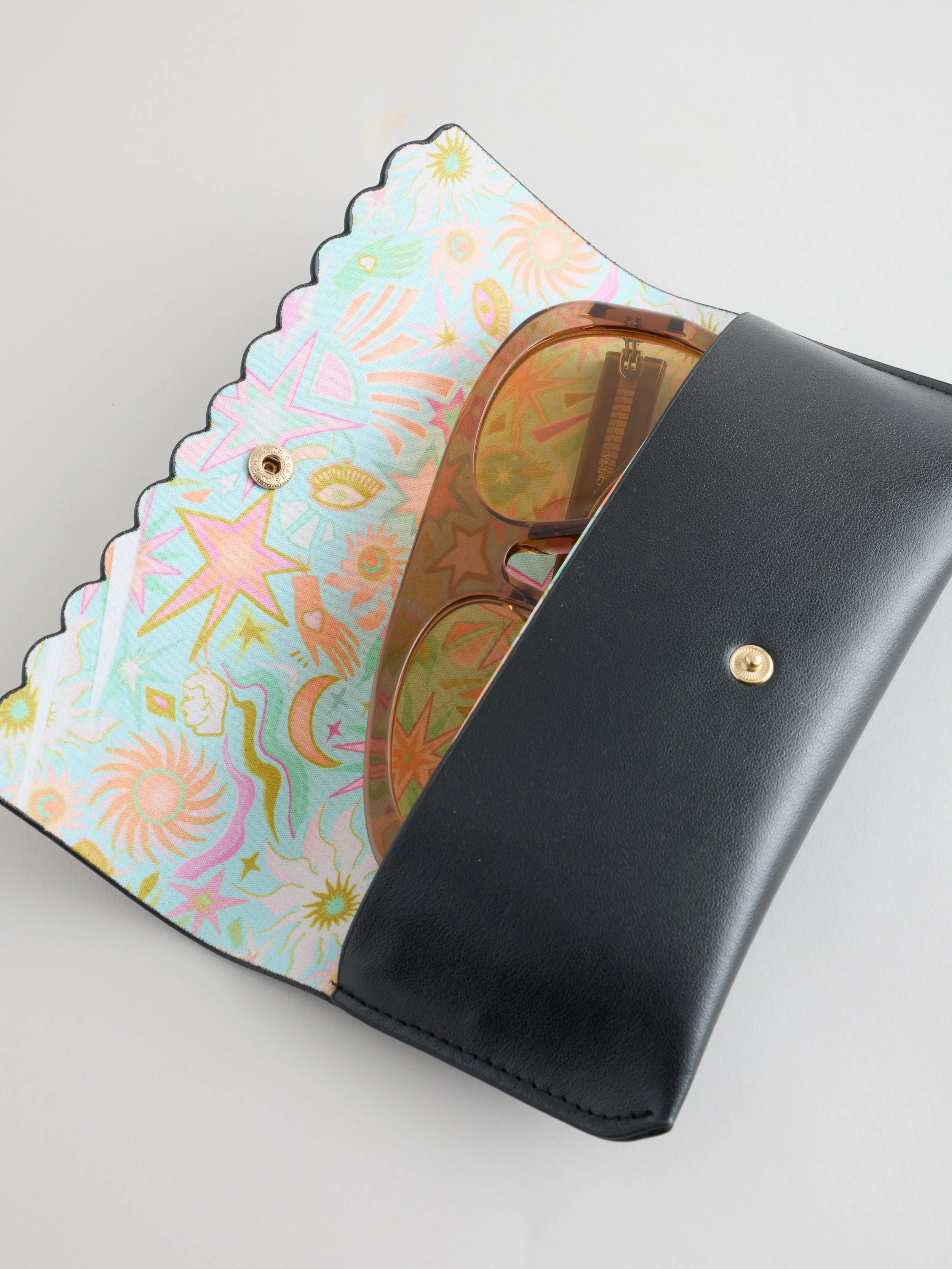 A black clutch purse with a flap that has a colorful, pastel pattern of stars, moons, and eyes, featuring a gold snap button closure, resting on a light gray surface.