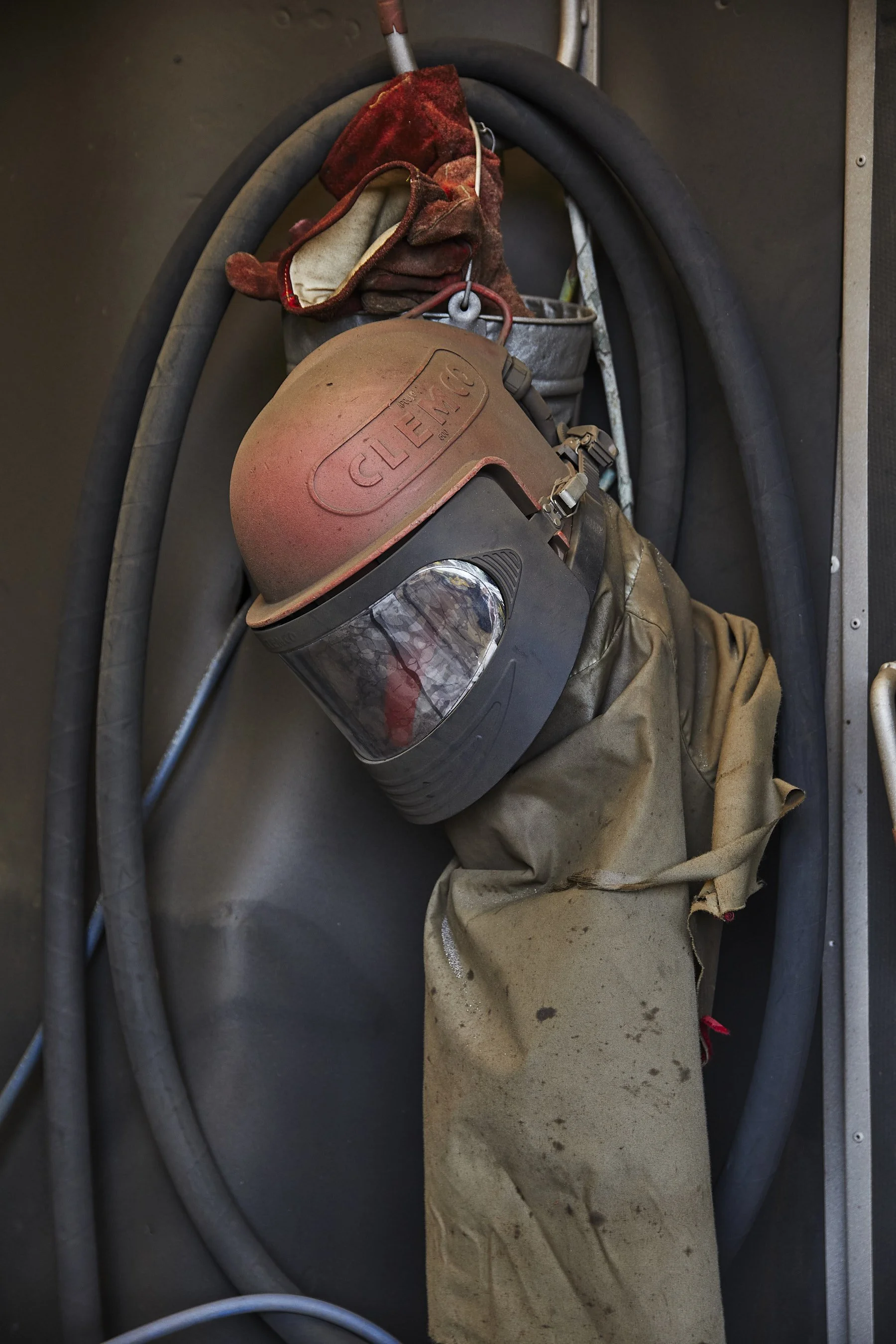 A firefighter's gear hanging on a wall, including a dusty hard hat with the brand name 'Cleveland,' a face shield, gloves, and a dirty cloth.