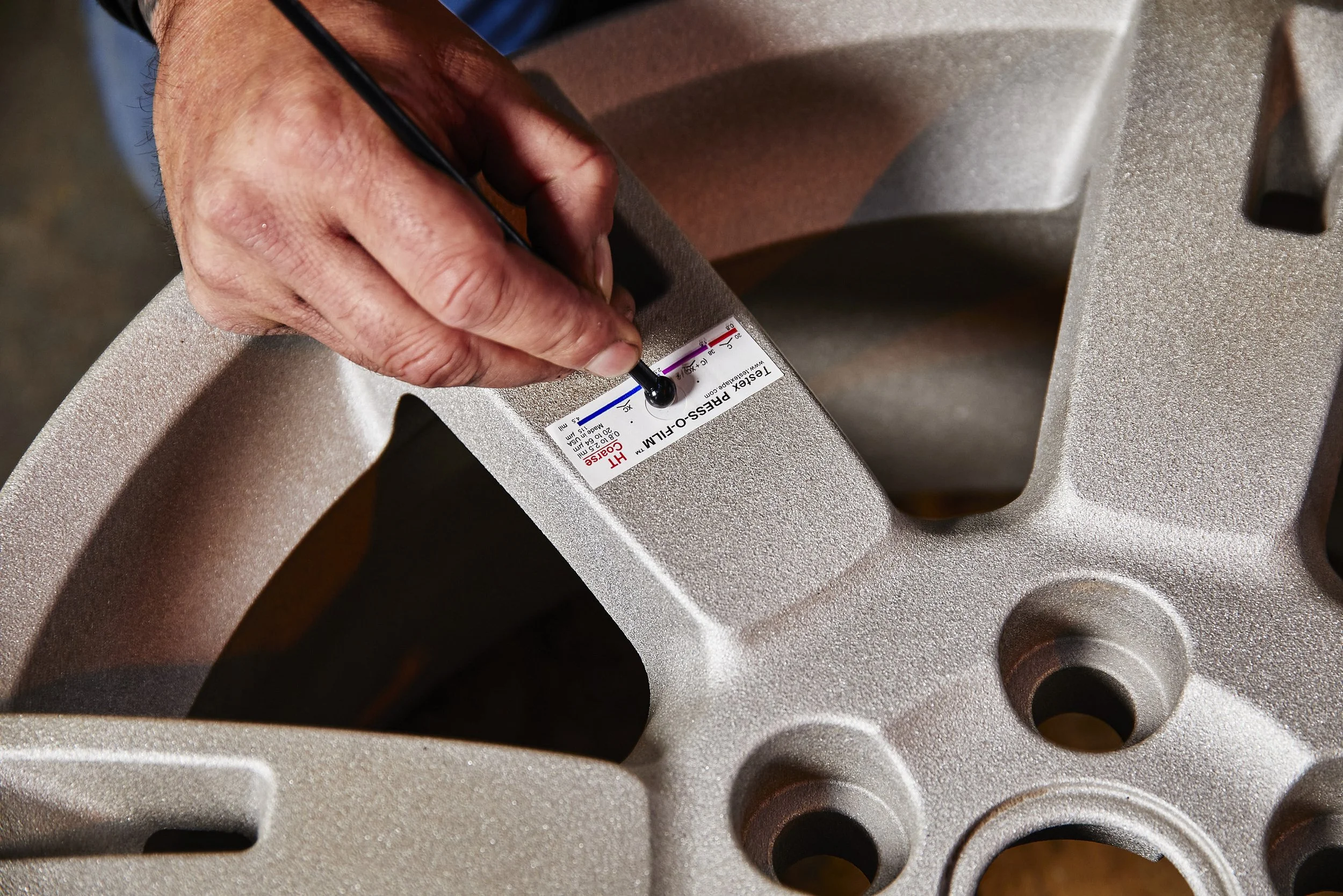 Close-up of someone using a black tool to adjust or test a metallic mechanical part with holes and a label that reads "TESTER PRESS-FILM."