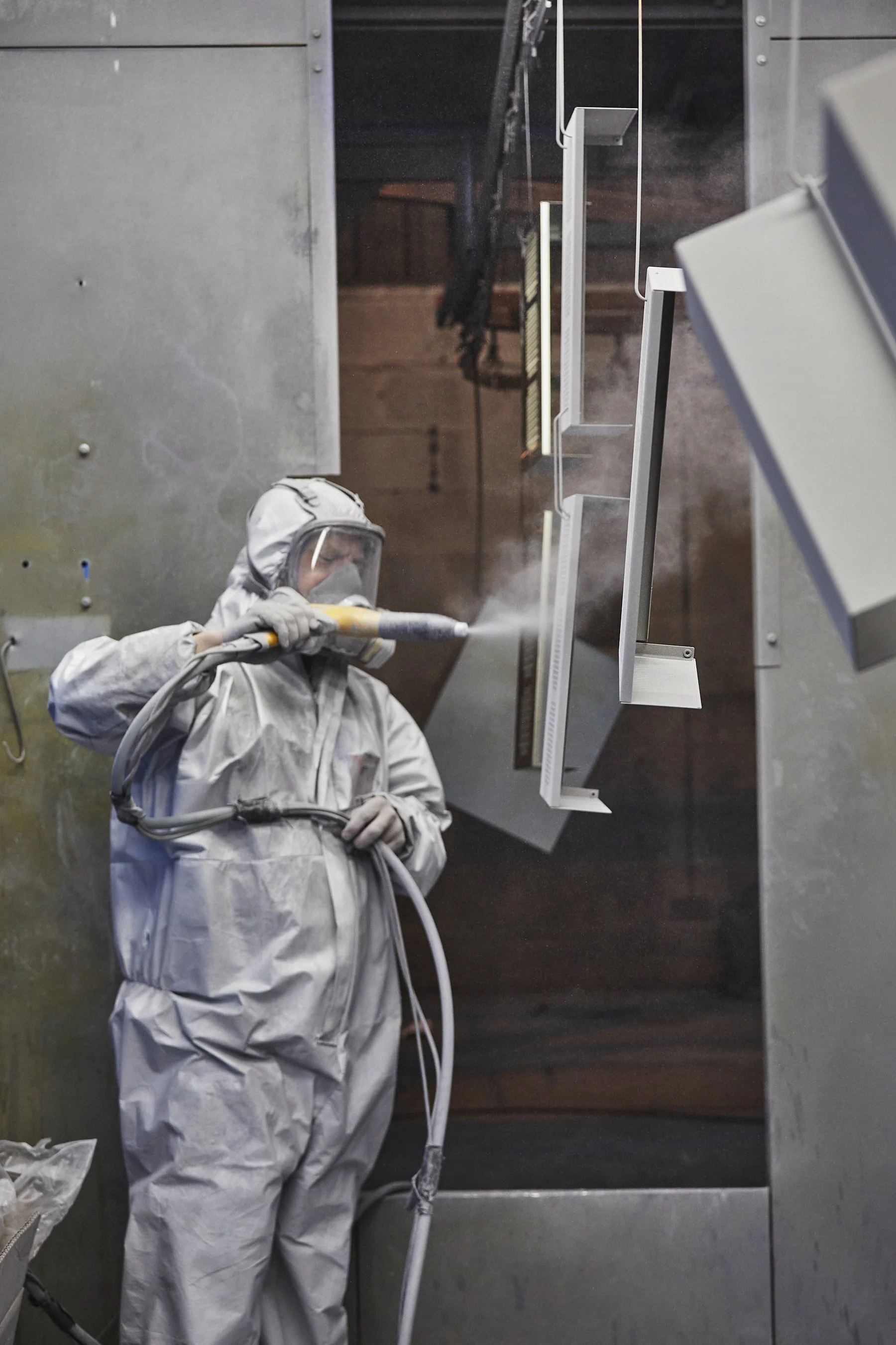 An individual wearing a white protective suit and mask is using a spray device in an industrial setting, with shelves and equipment in the background.