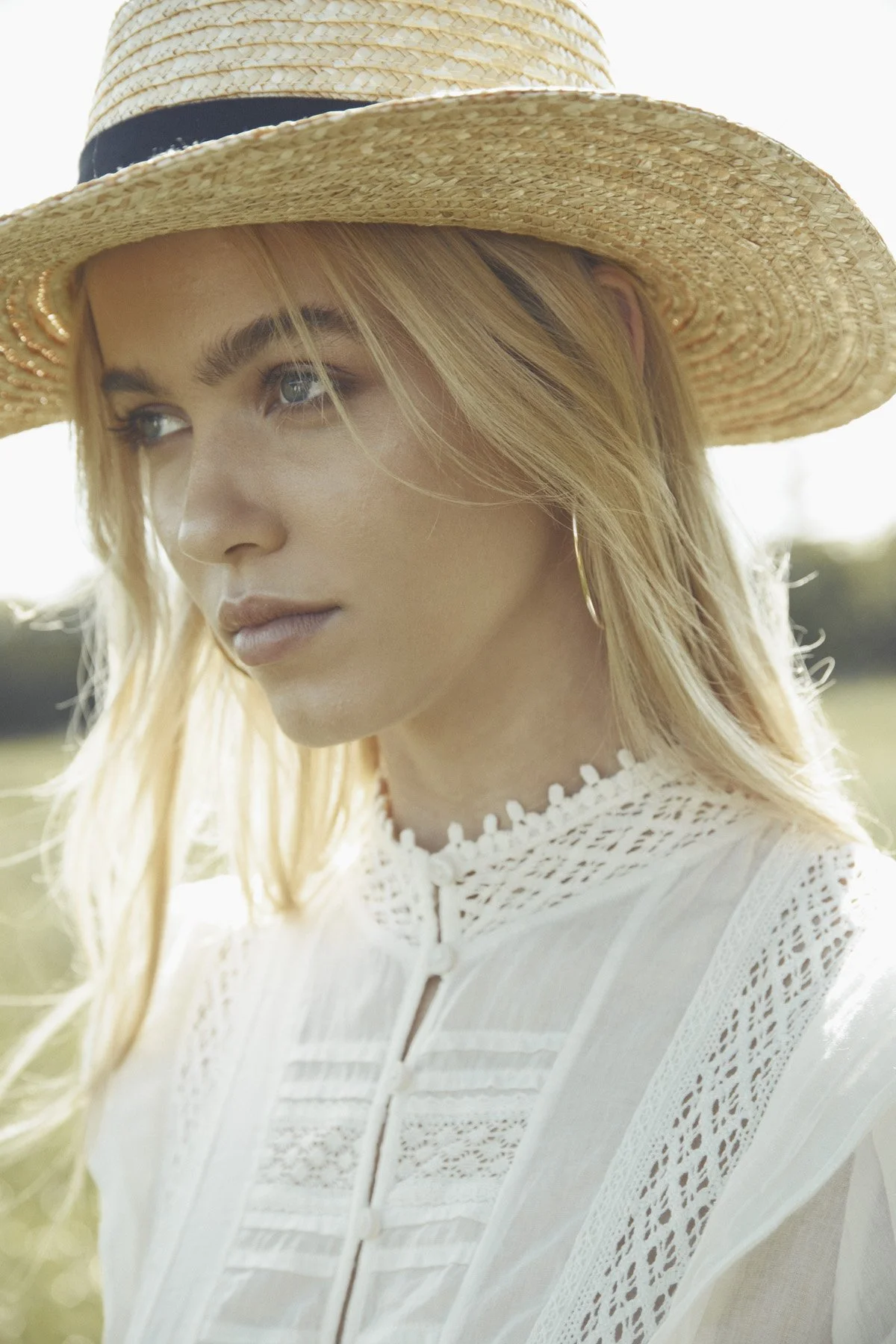 A close-up of a young woman with blonde hair wearing a wide straw hat with a black band and a white lace dress, standing outdoors with sunlight.