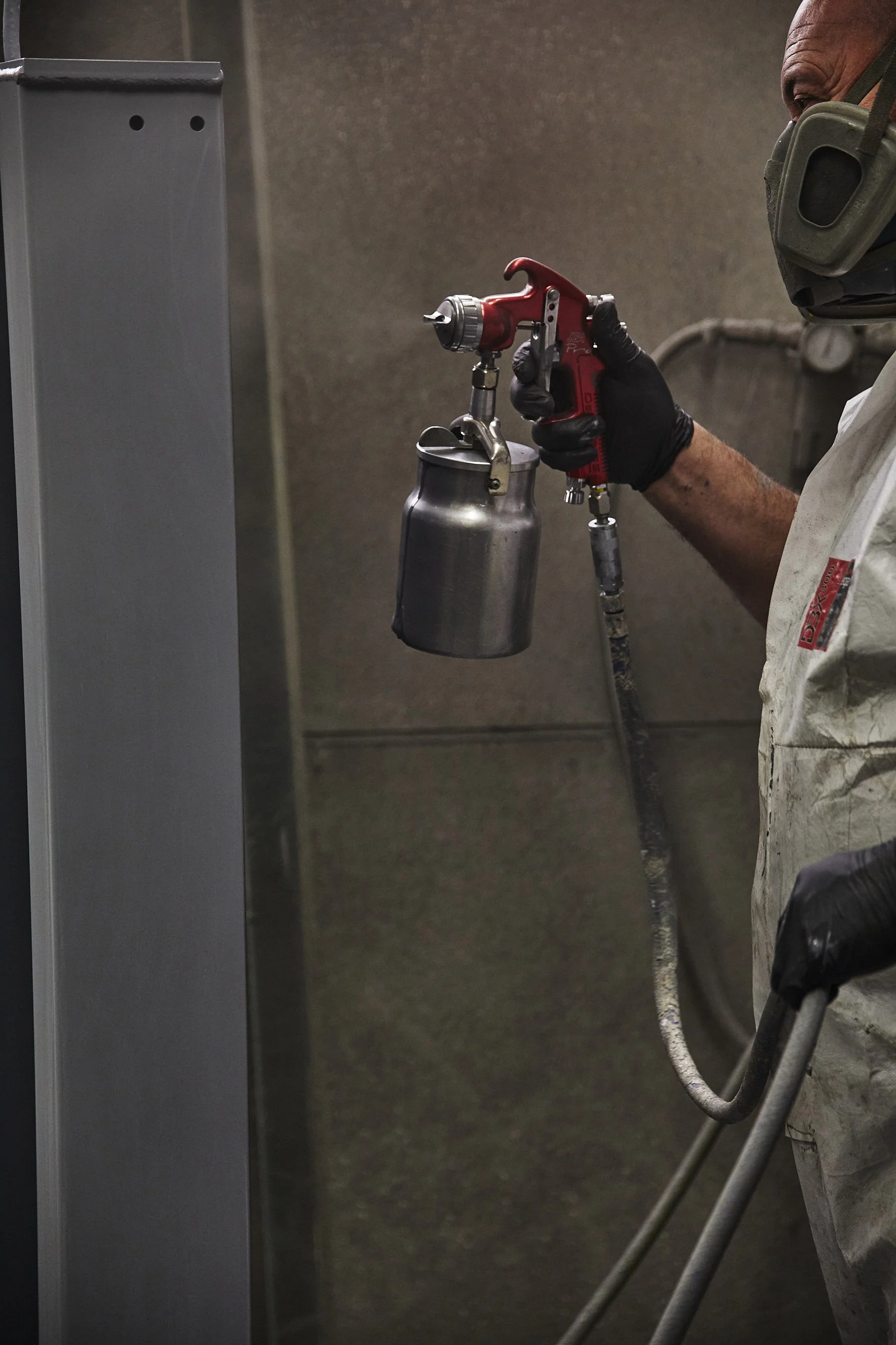 A person wearing black gloves and safety gear using a spray gun to paint a metal surface in a workshop or paint booth.