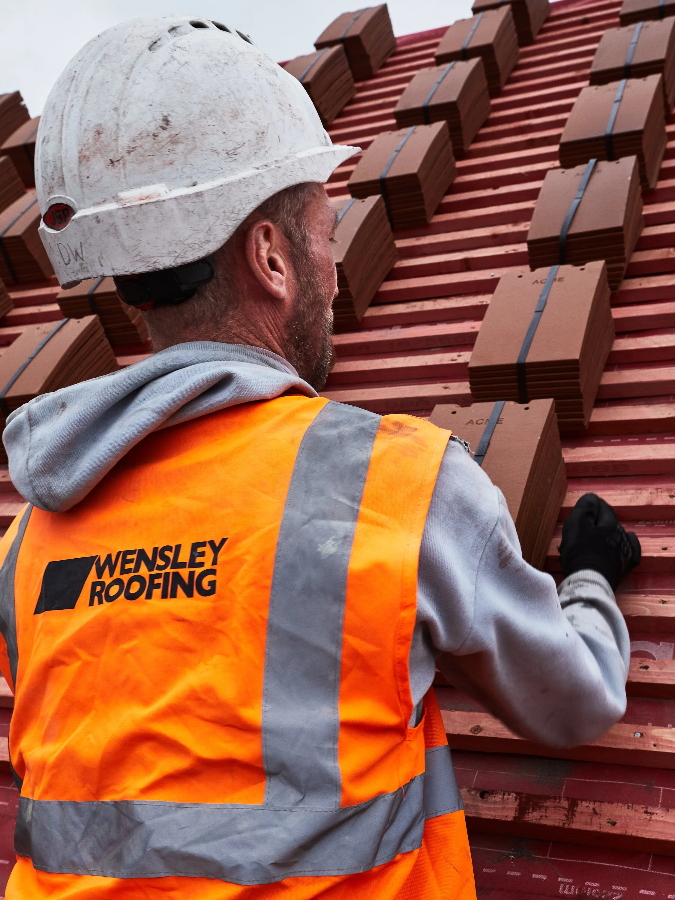 A construction worker wearing a white hard hat and an orange safety vest with "Wensley Roofing" printed on the back, working on a roof with stacked tiles.