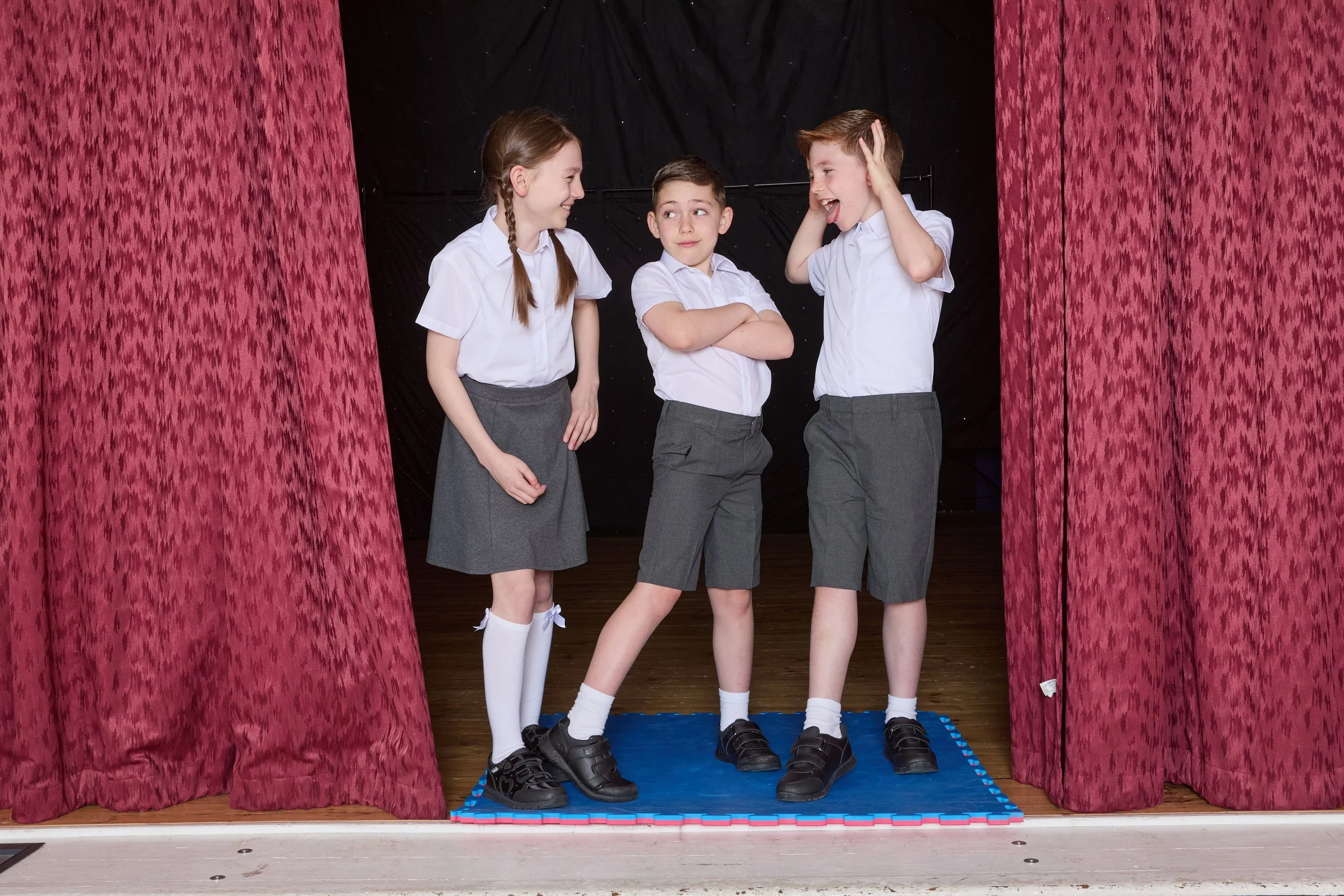 Three children in school uniforms standing on a small blue mat backstage between red curtains, smiling and making faces at each other.