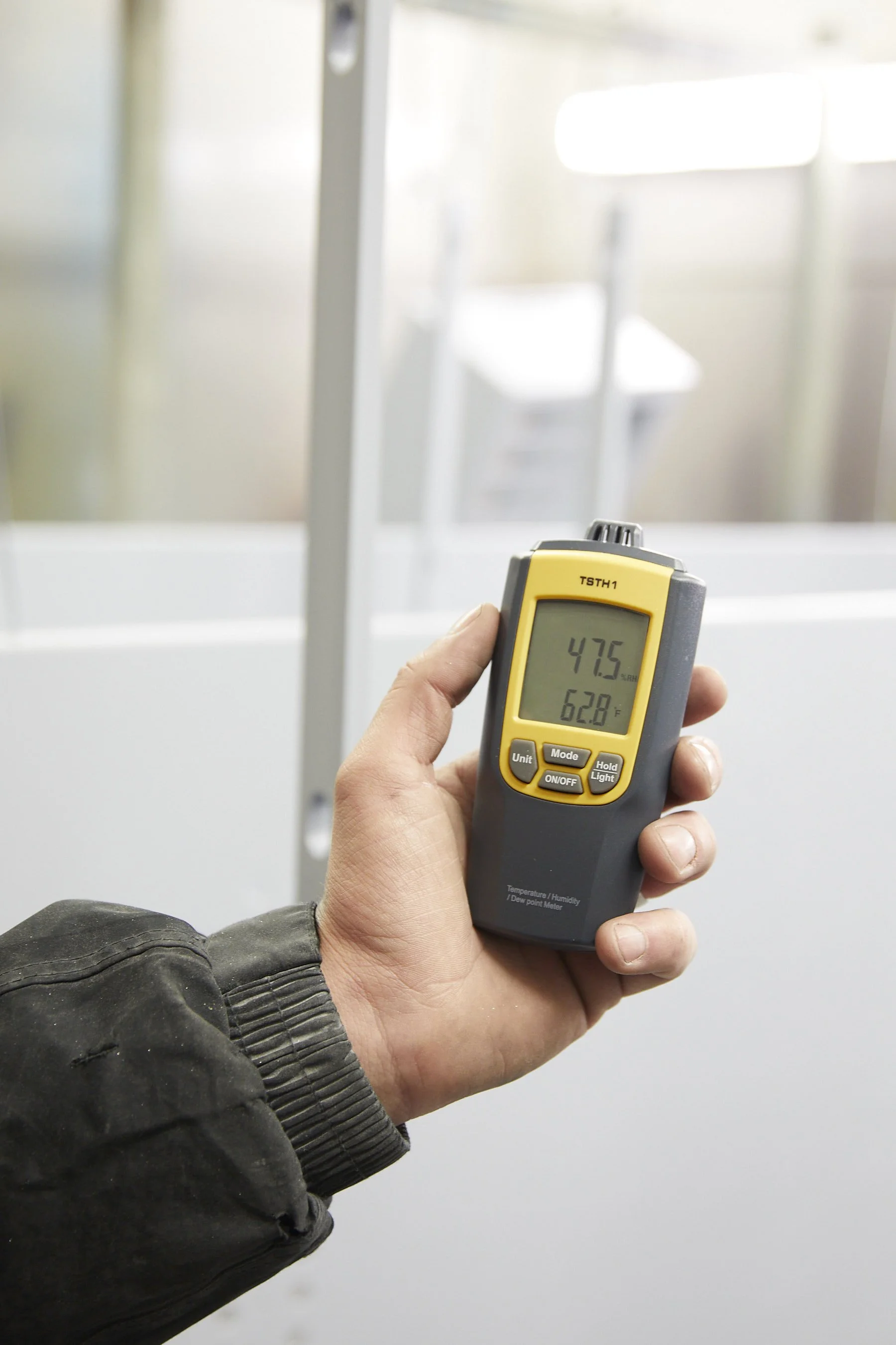 A person holding a device displaying a temperature reading of 47.5°F, with a blurred background of an indoor or industrial setting.