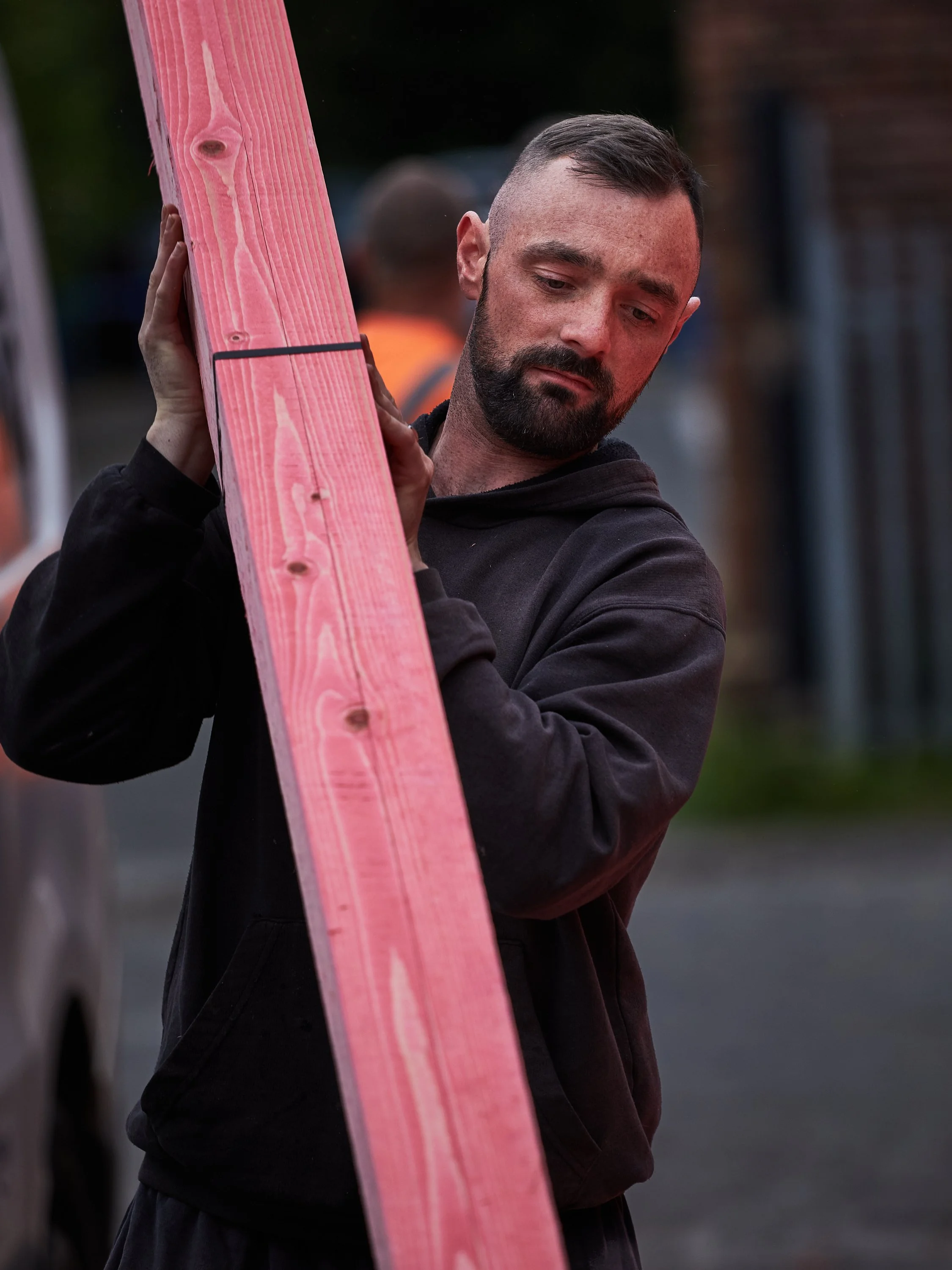 A man with a beard and short hair carrying a piece of pink construction lumber on his shoulder