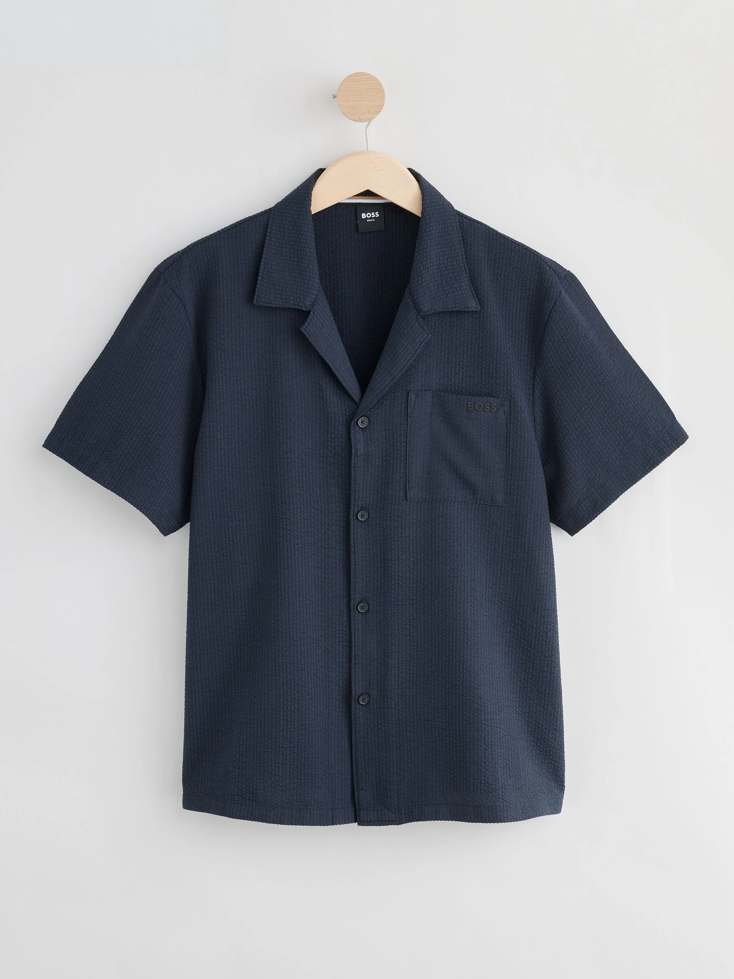 A navy blue short-sleeve button-up shirt hanging on a wooden hanger against a plain white wall.