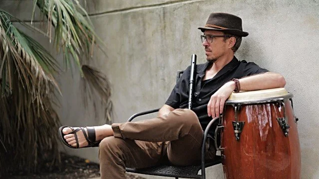 Man sitting outdoors near a concrete wall with a conga drum, wearing a hat, sunglasses, black shirt, and brown pants.