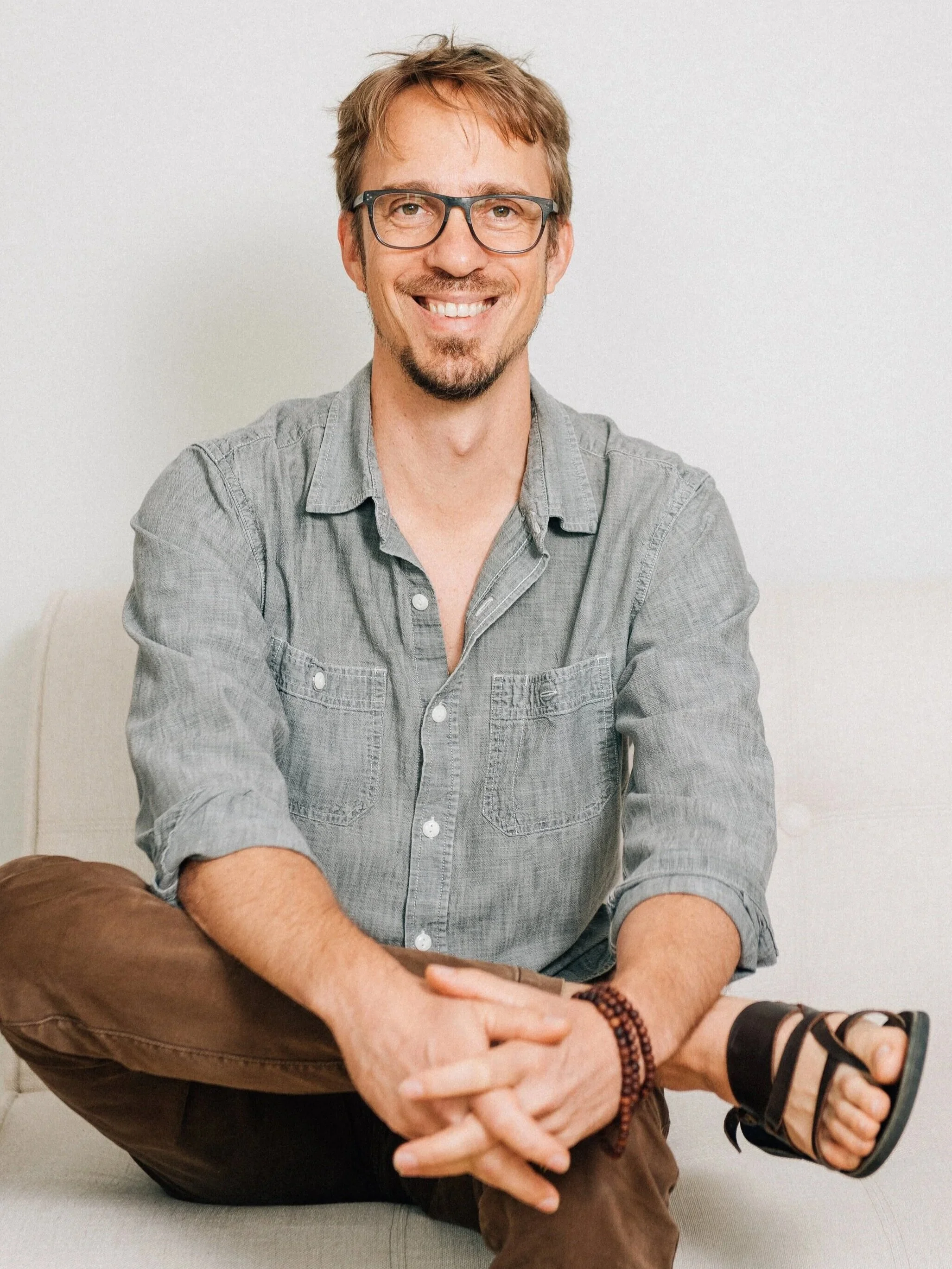 A man with glasses, a beard, and light brown hair, wearing a gray button-up shirt and brown pants, sitting cross-legged on a light-colored couch against a plain white wall, smiling at the camera.