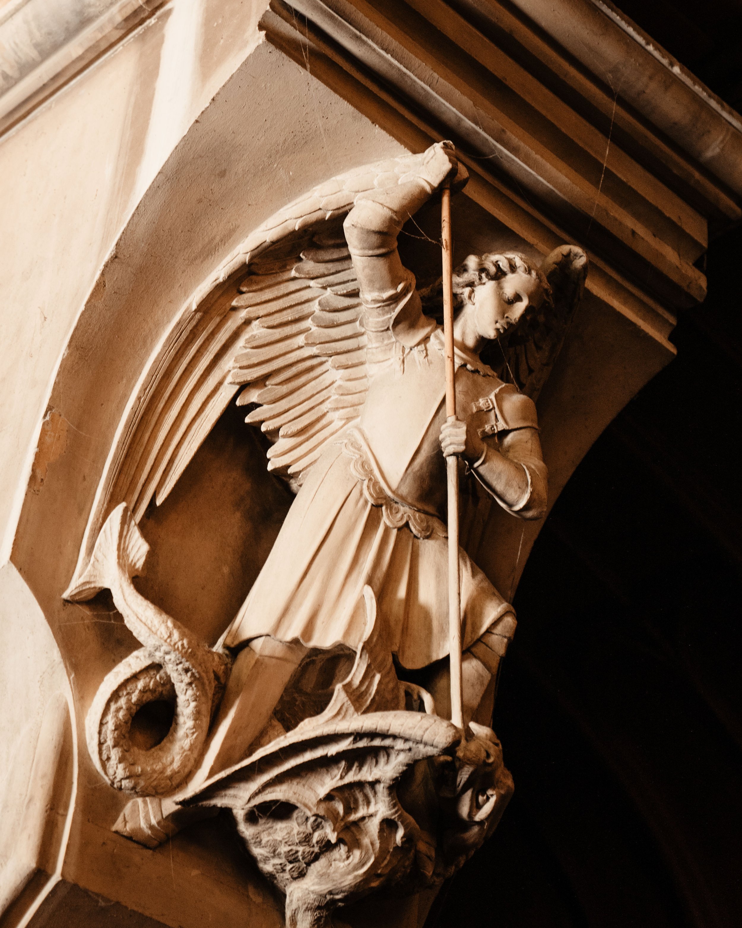Stone sculpture of an angel with wings, holding a spear, standing over a dragon-like creature in a classical architectural niche.