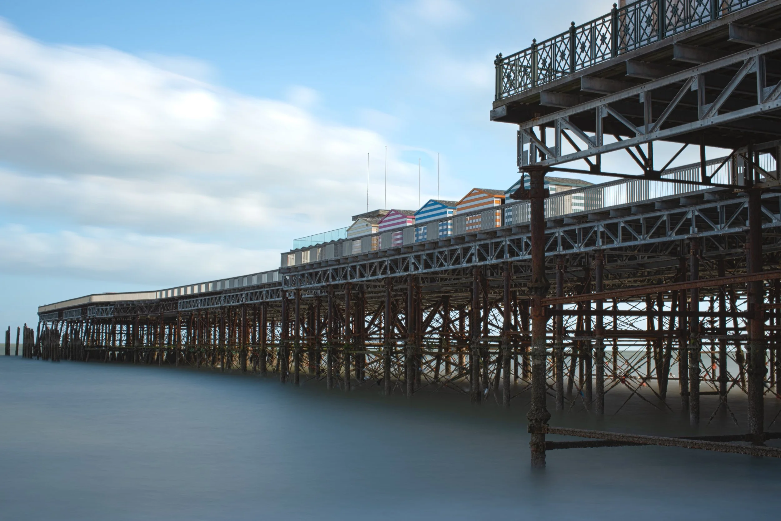 A traditional seaside pier with colorful beach huts, metal structure, calm water, and cloudy sky.