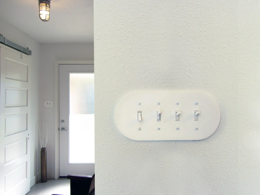 Installed plodes Rounded Series wall plates: an entryway view beside a close-up of a white four-gang toggle switch plate on a textured wall