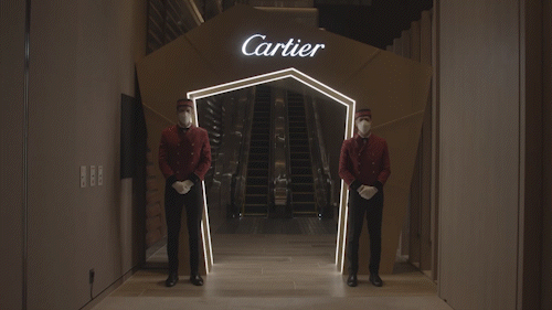 Two staff members wearing masks and red uniforms standing in front of a Cartier store entrance with escalators inside.