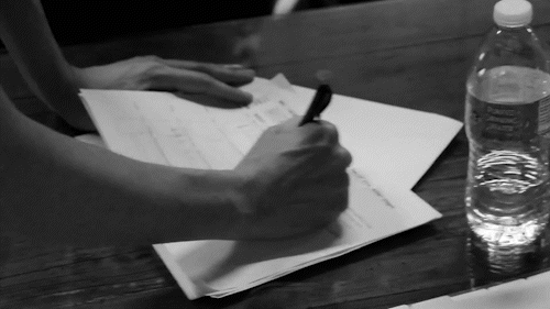 Person's hand writing with a pen on papers, with a water bottle on a wooden table.