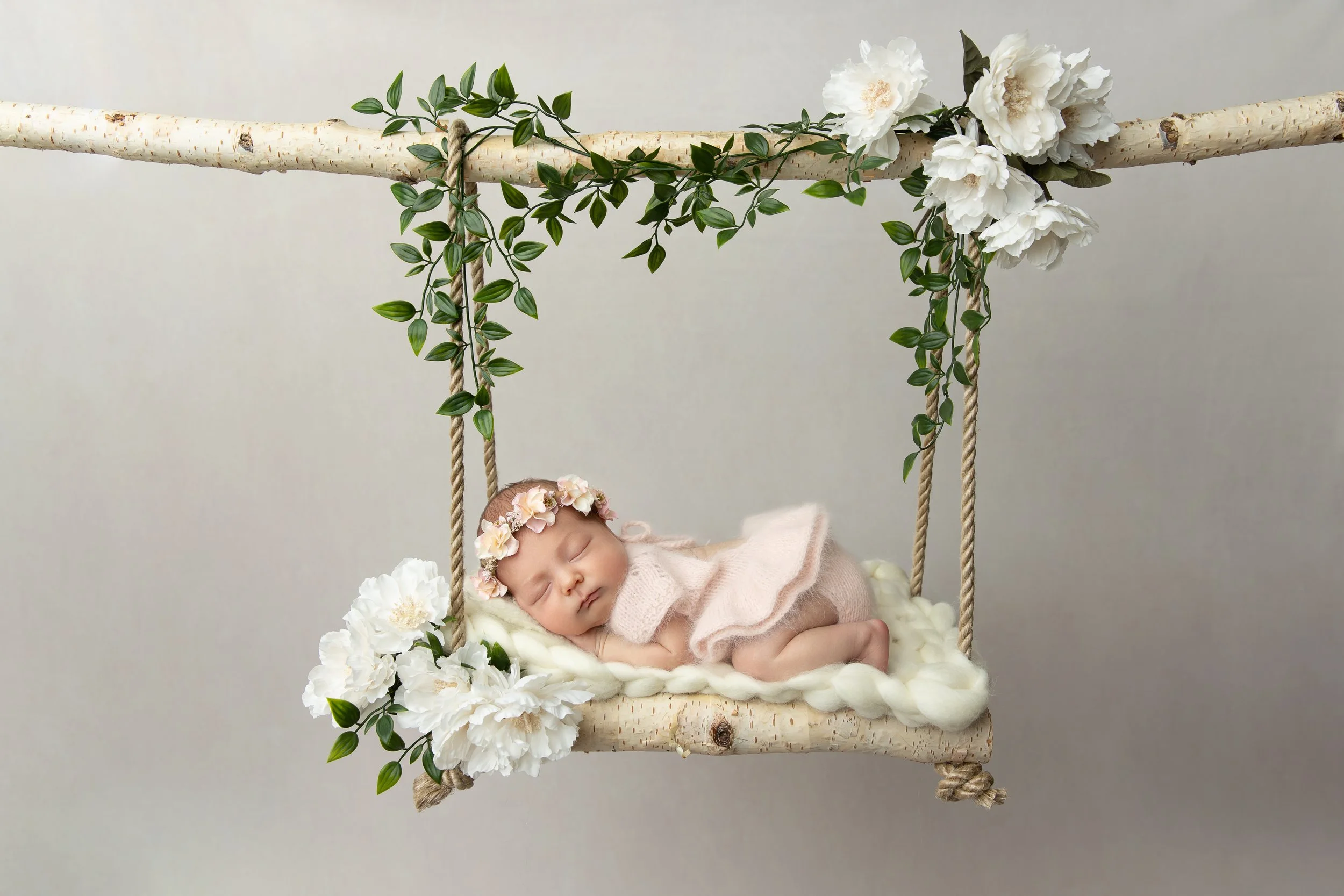 Baby sleeping on a swing decorated with white flowers and green leaves.