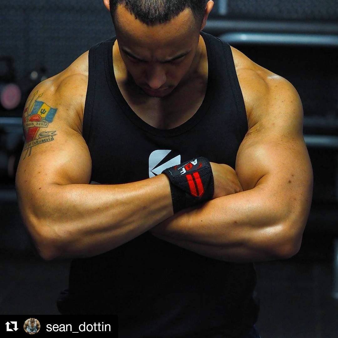Bodybuilder with muscular arms and tattoo, wearing a black tank top and workout gloves, arms crossed.